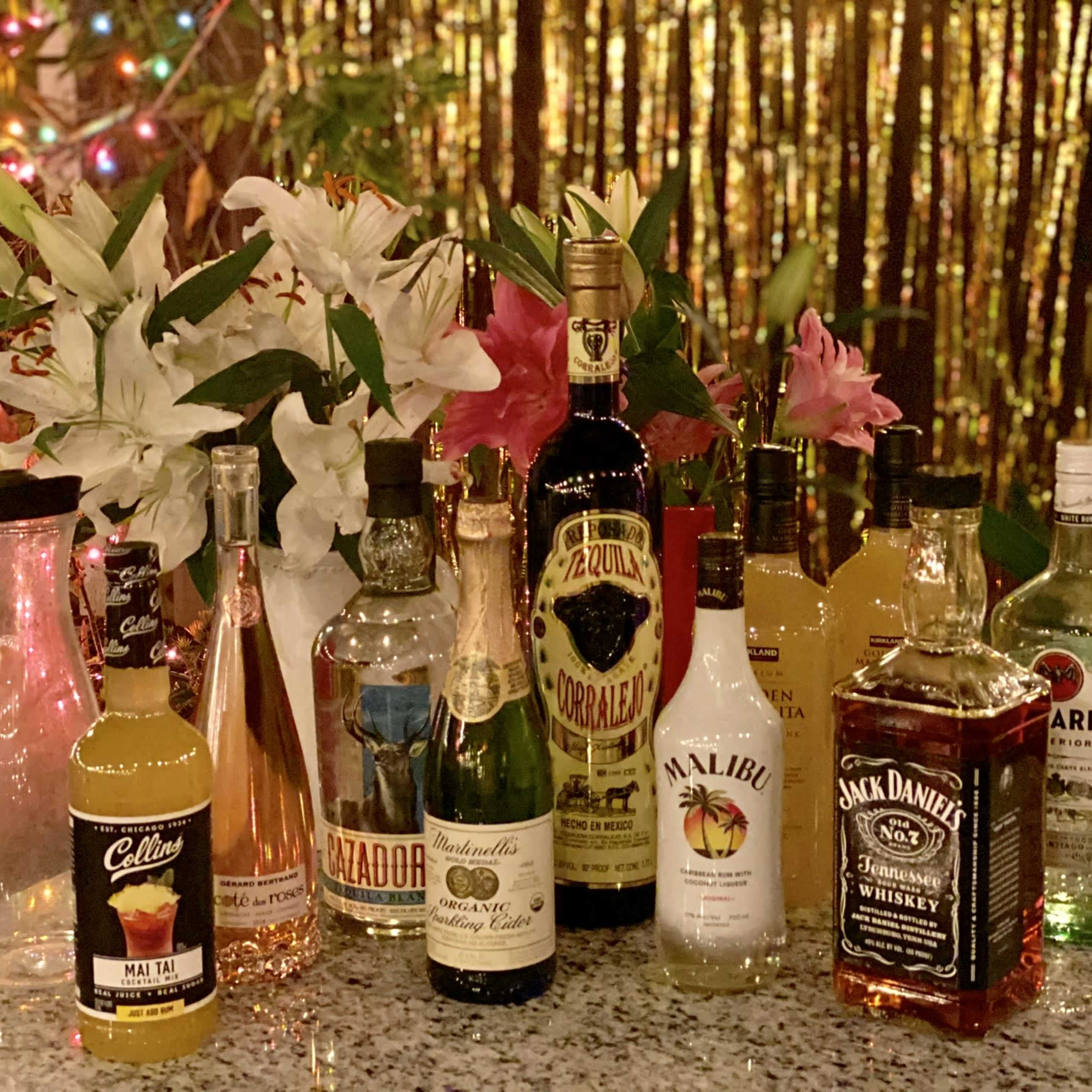 A variety of alcoholic drinks arranged on a table alongside decorative flowers, with a festive backdrop of lights.