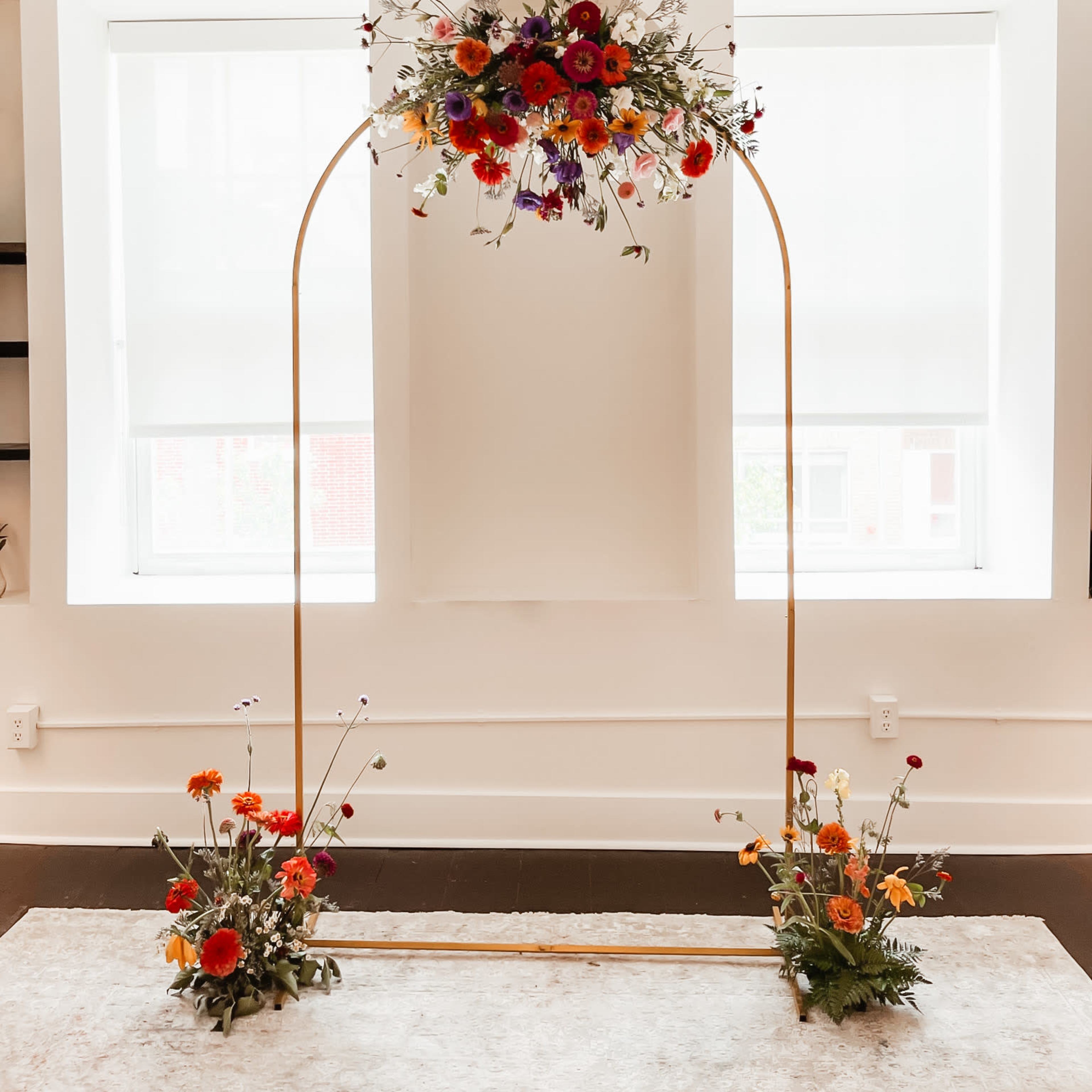 A floral arch made of metal, adorned with colorful flowers, positioned between two windows in a bright room.