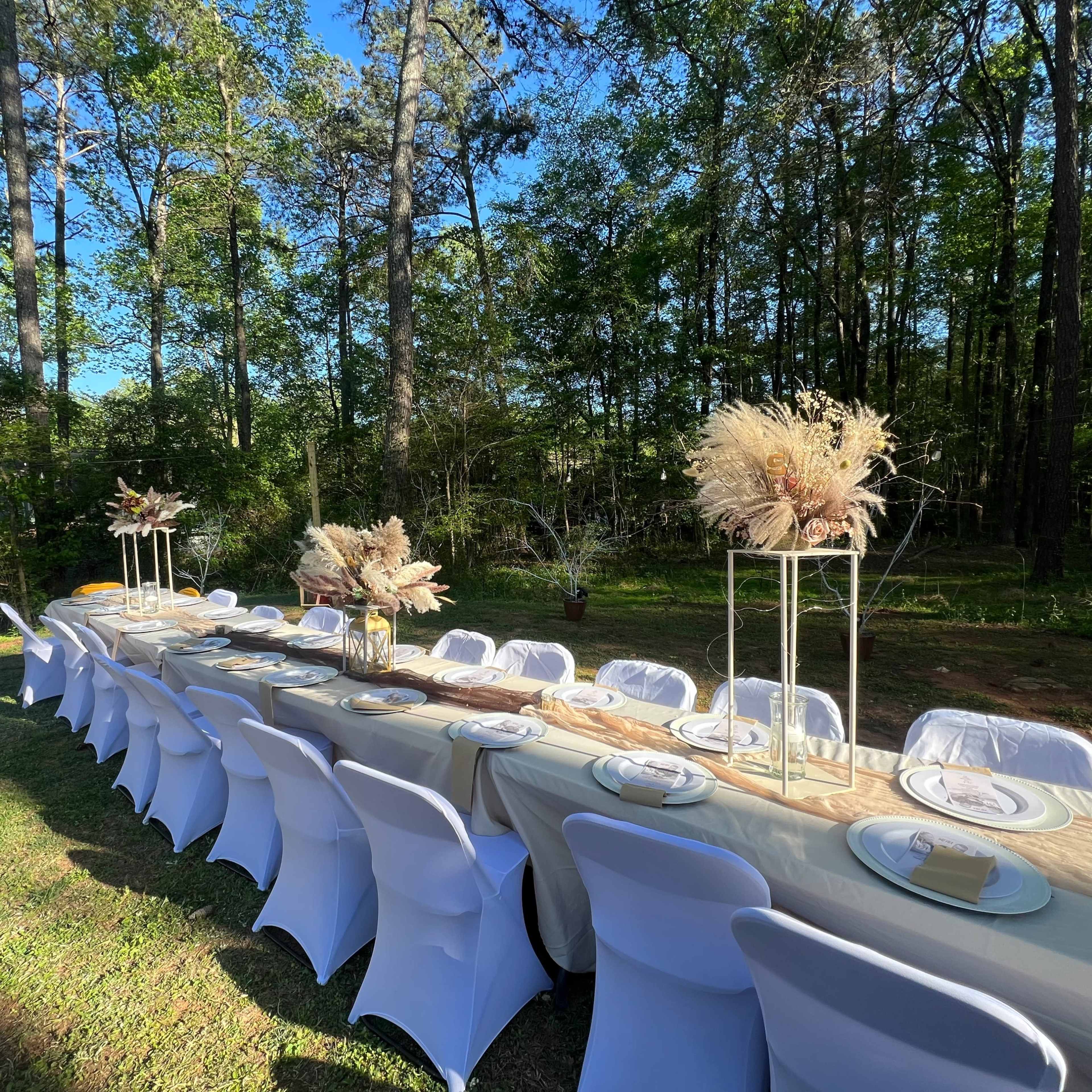 A long outdoor dining table is set with decor and chairs amidst a wooded area.