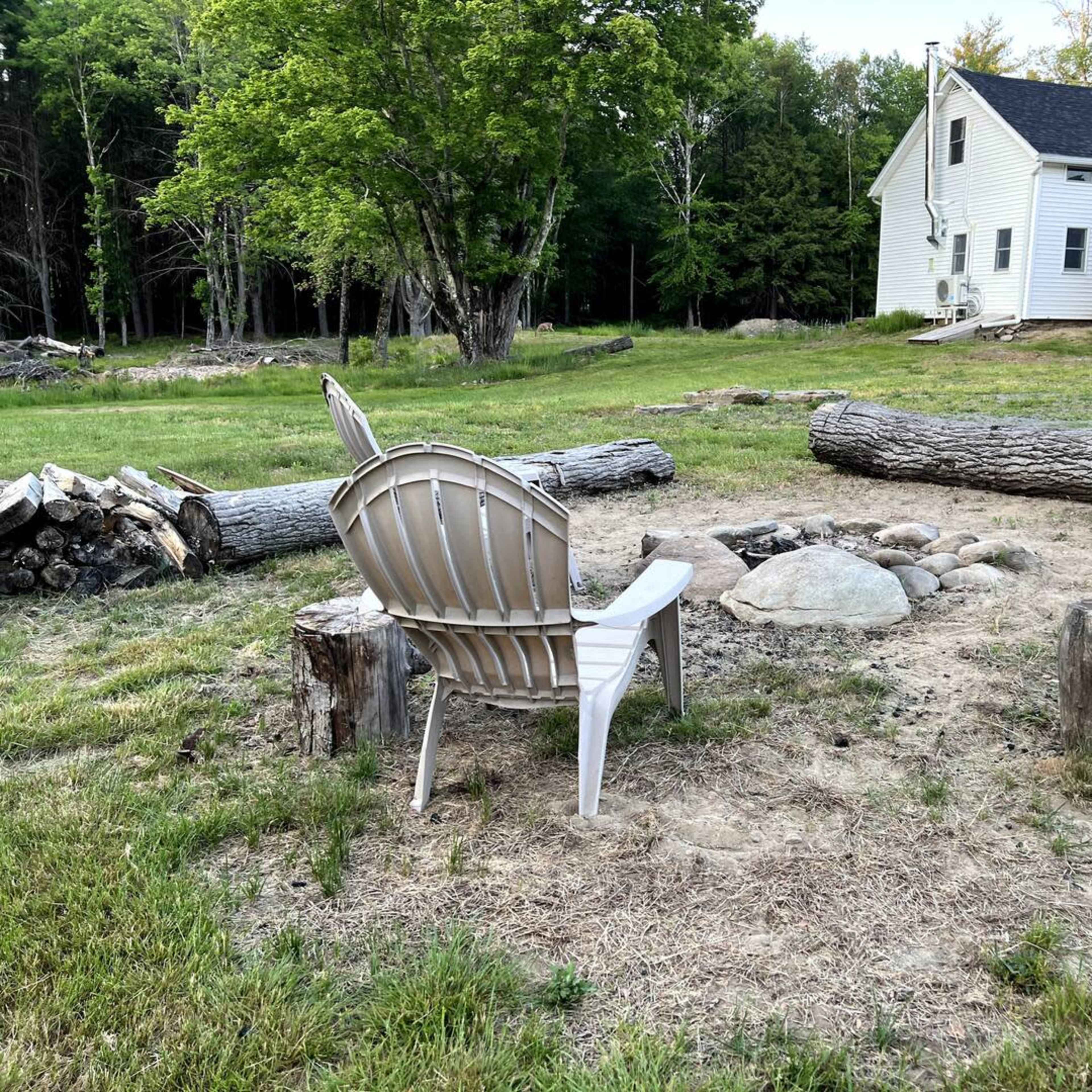 A single plastic chair sits beside a fire pit made of stones, surrounded by logs and a grassy area next to a weathered house in a forested setting.