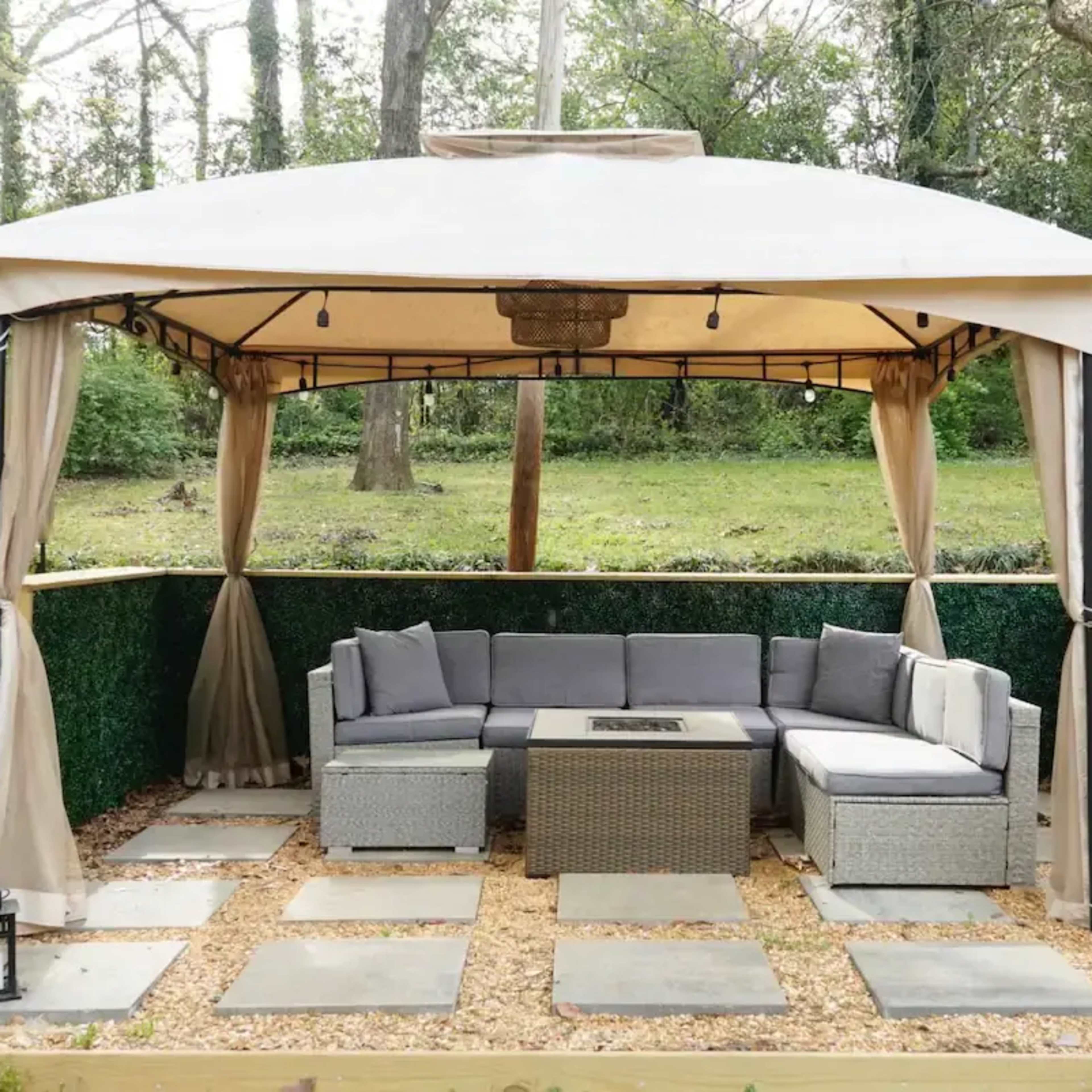 A gazebo with a sectional seating area and fire pit is situated on a gravel patio surrounded by greenery.