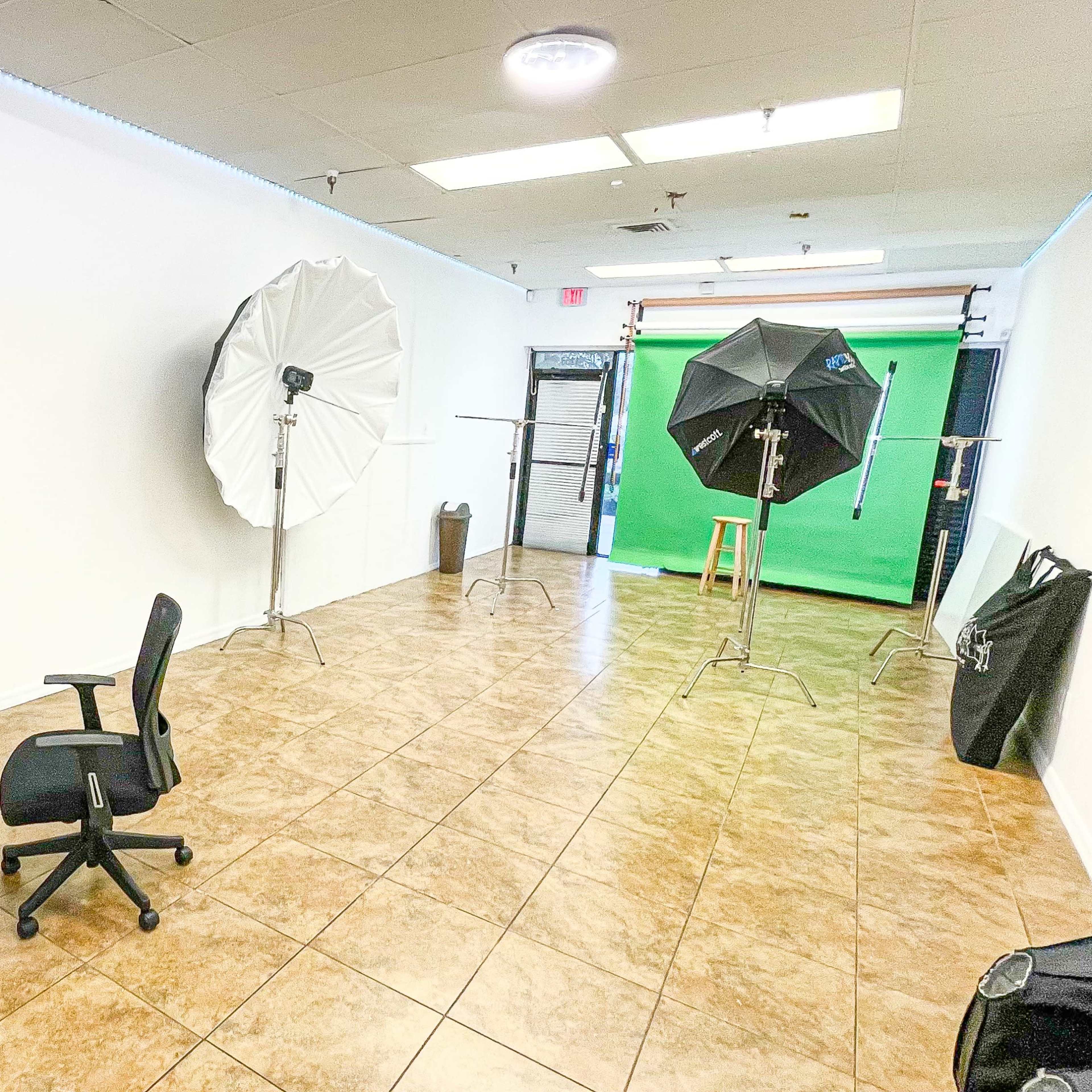 A studio space with a green screen, two lighting softboxes, a computer desk with an office chair, and tiled flooring.