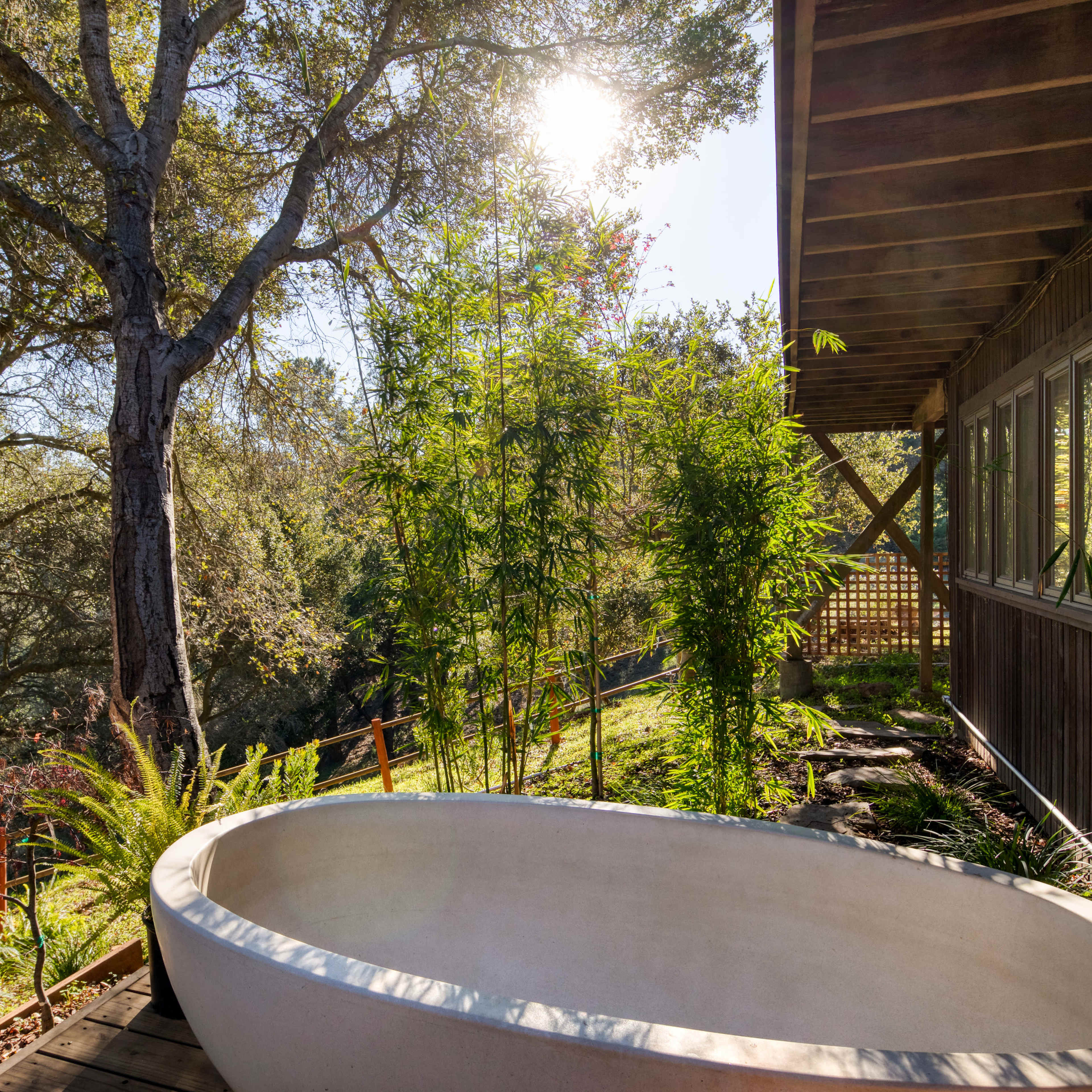 A freestanding white bathtub is positioned on a wooden deck surrounded by plants and trees, with sunlight filtering through the branches.