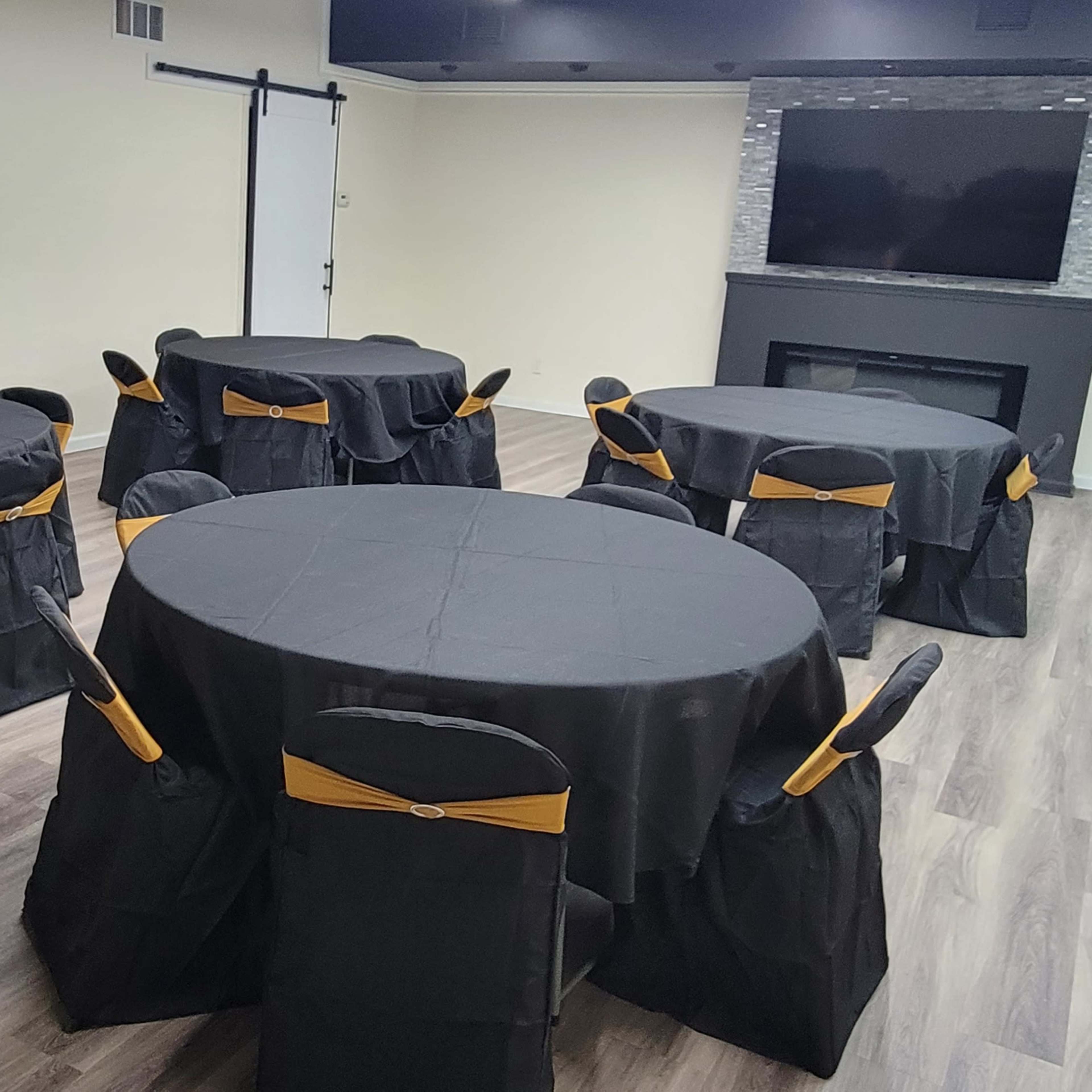 A room set up for an event, featuring several round tables covered with black tablecloths and decorated with gold chair sashes.