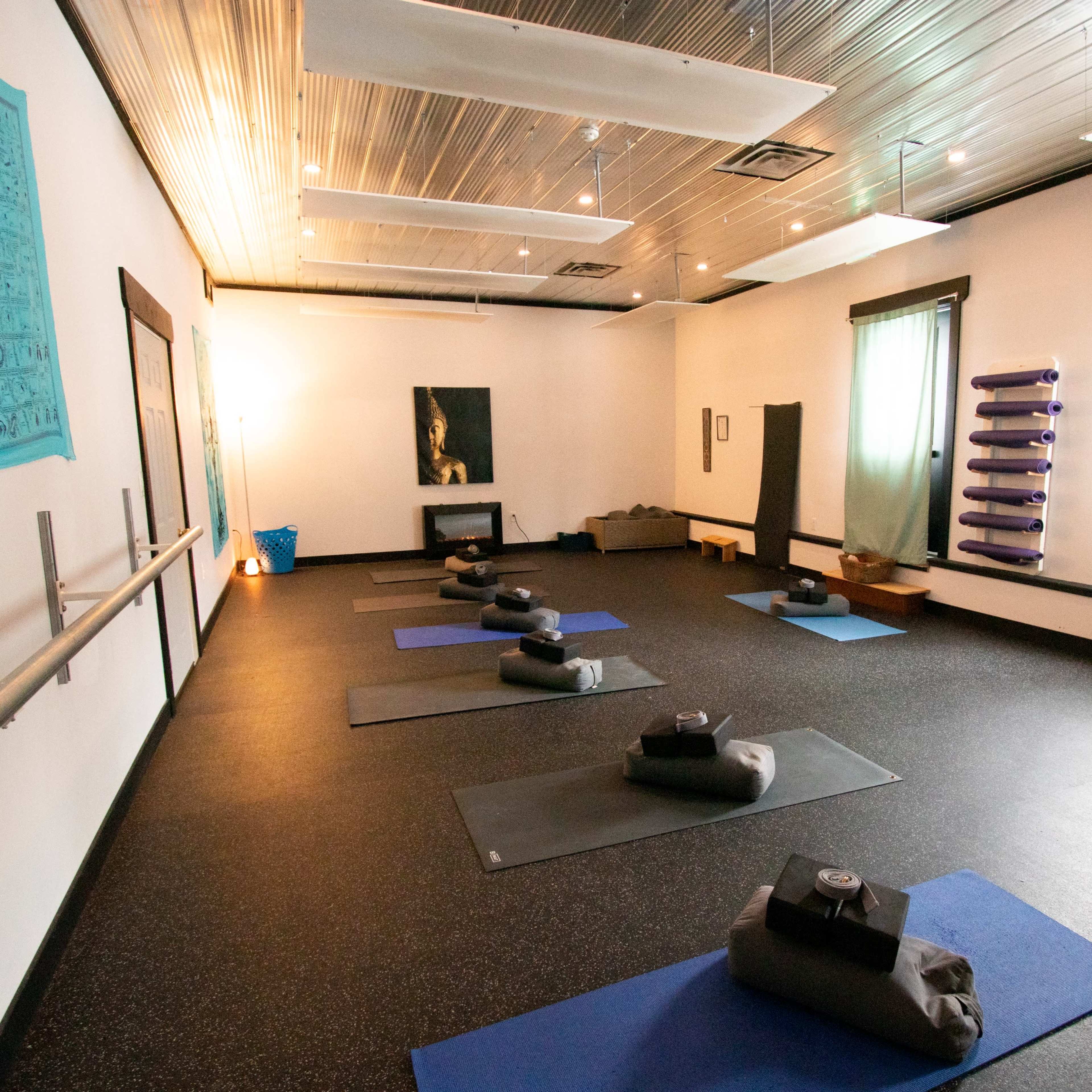 A spacious yoga studio is arranged with mats and bolsters organized neatly on a dark floor, complemented by soft lighting and simple decor.