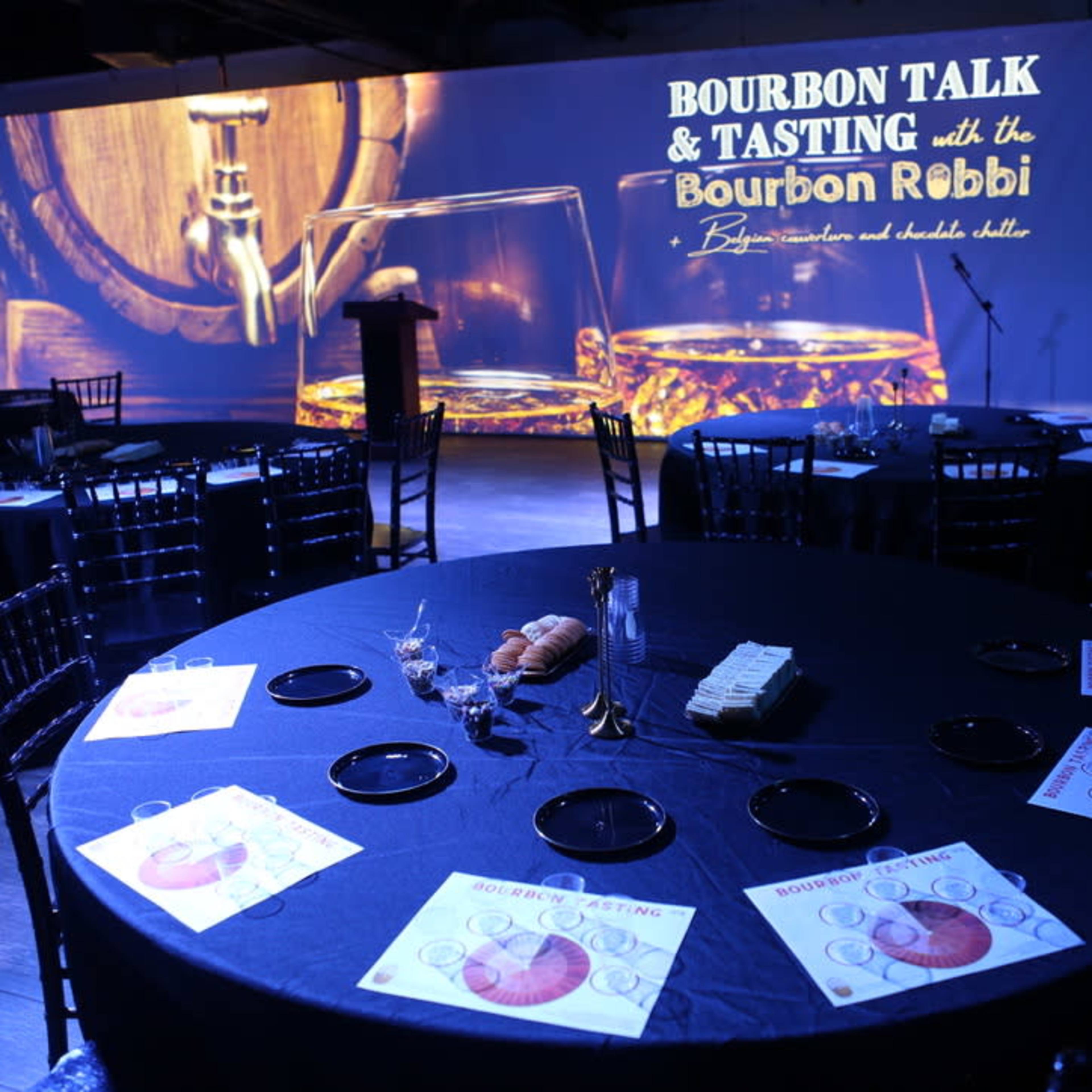 A round table is set with tasting materials and snacks in a dimly lit room featuring a large screen displaying "Bourbon Talk & Tasting."