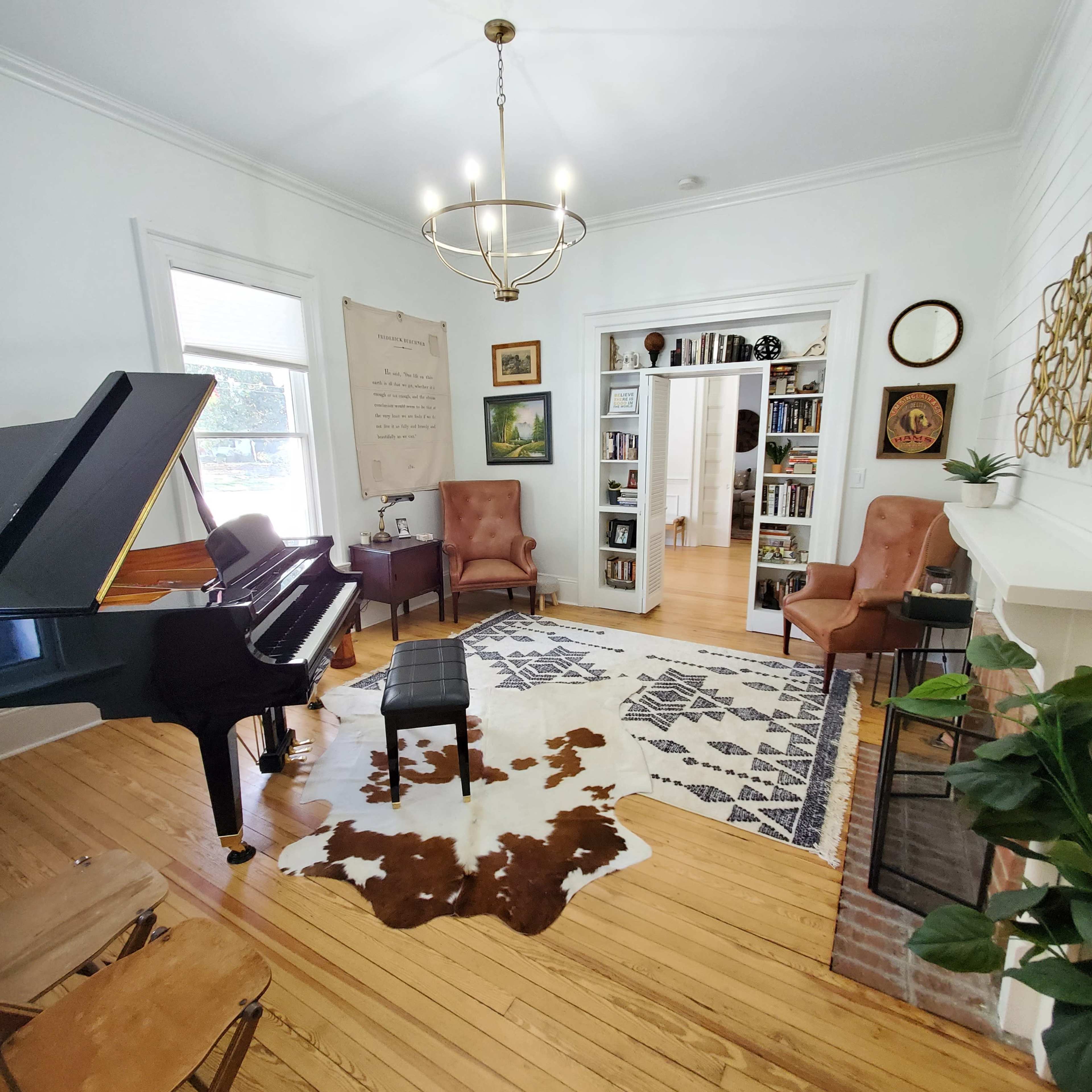 A bright living room features a black piano, two brown chairs, a patterned rug, and a bookshelf in the background.