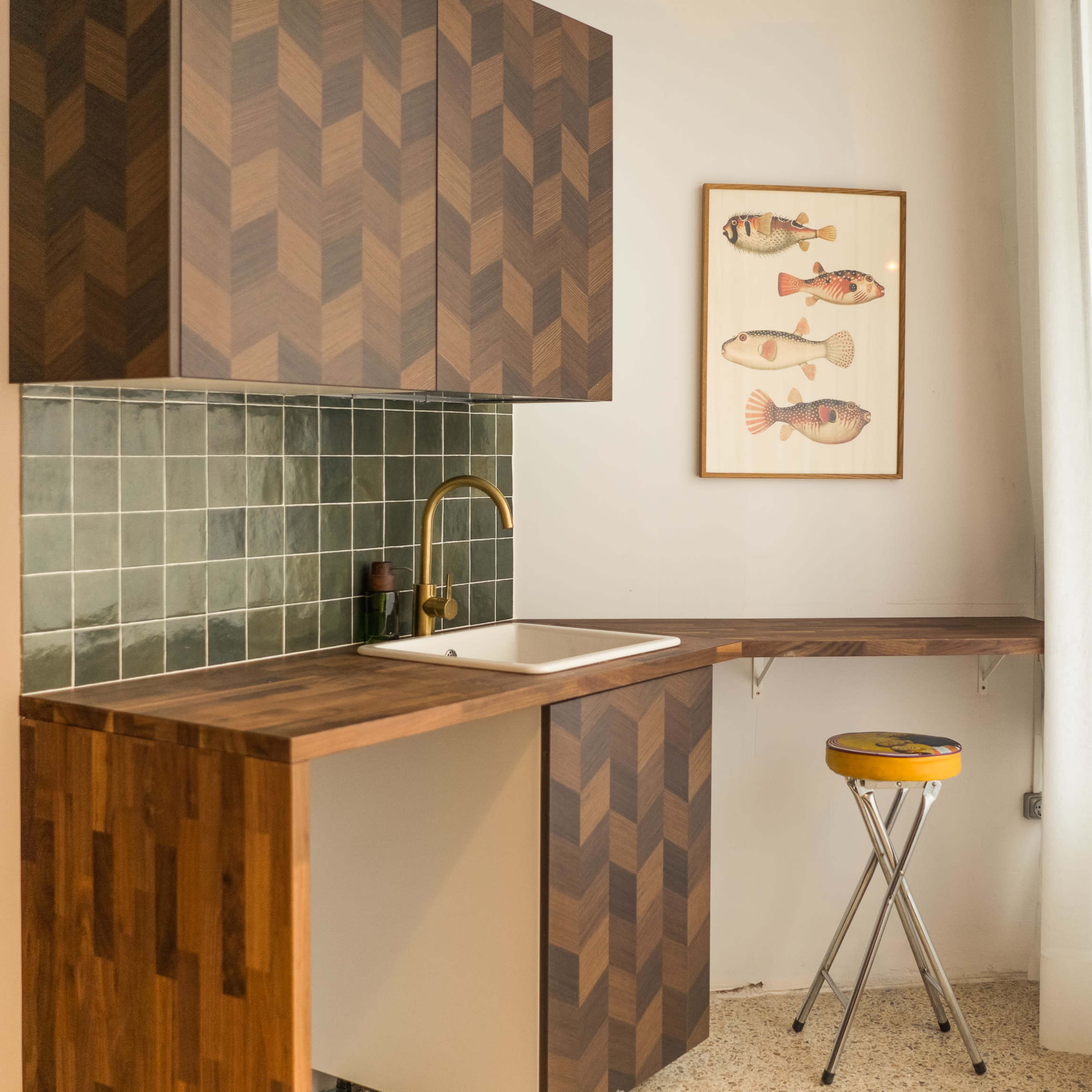 A corner kitchen with a wooden countertop, a white sink, green tiled backsplash, and a framed fish illustration on the wall.