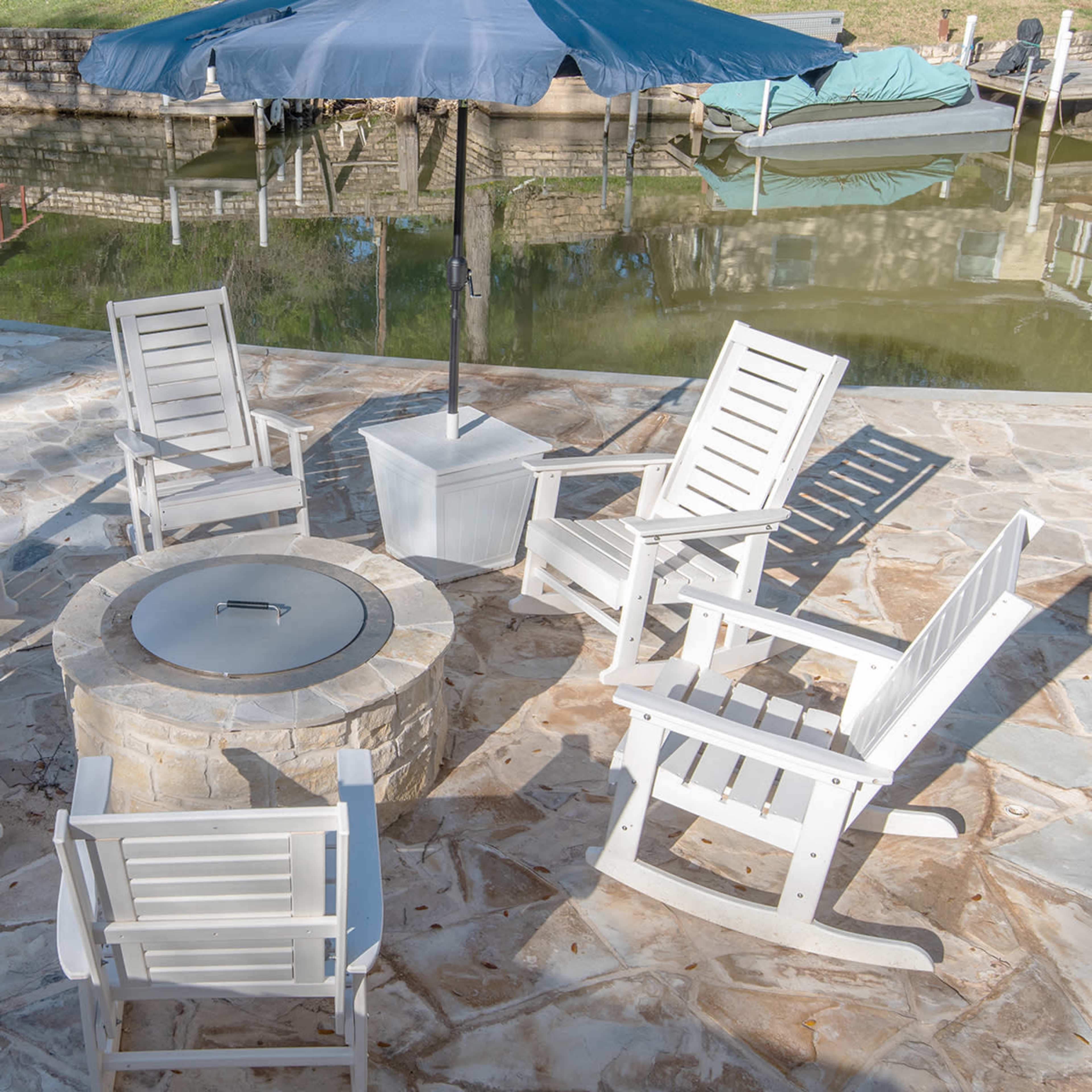 A circular arrangement of white rocking chairs surrounds a stone fire pit on a paved patio beside a body of water.