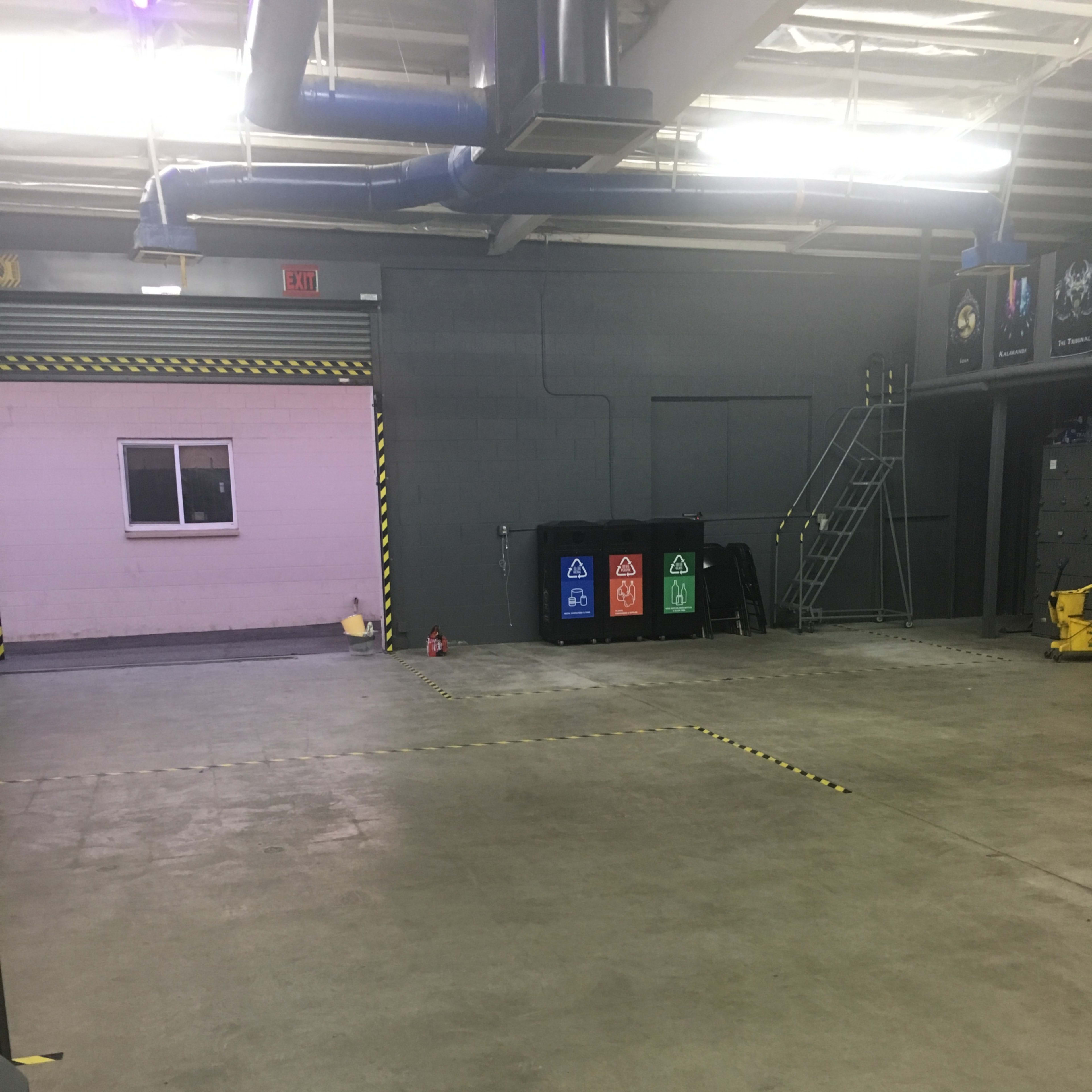 A large, empty warehouse or garage space with a concrete floor, a small window on the back wall, and recycling bins in three colors lined up against the wall.
