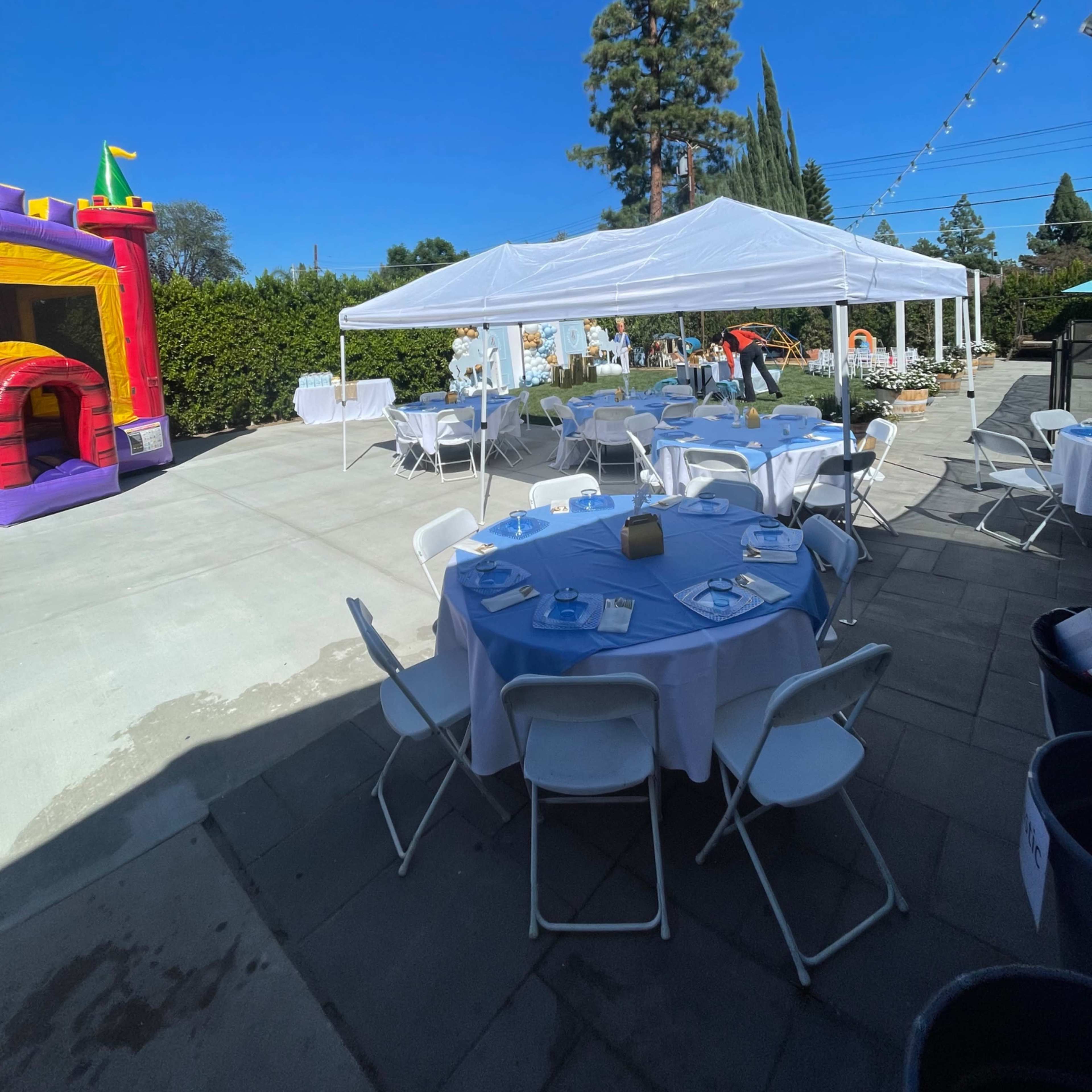 A tent-covered area with round tables and white chairs is set up near a bouncy castle and greenery under a clear blue sky.