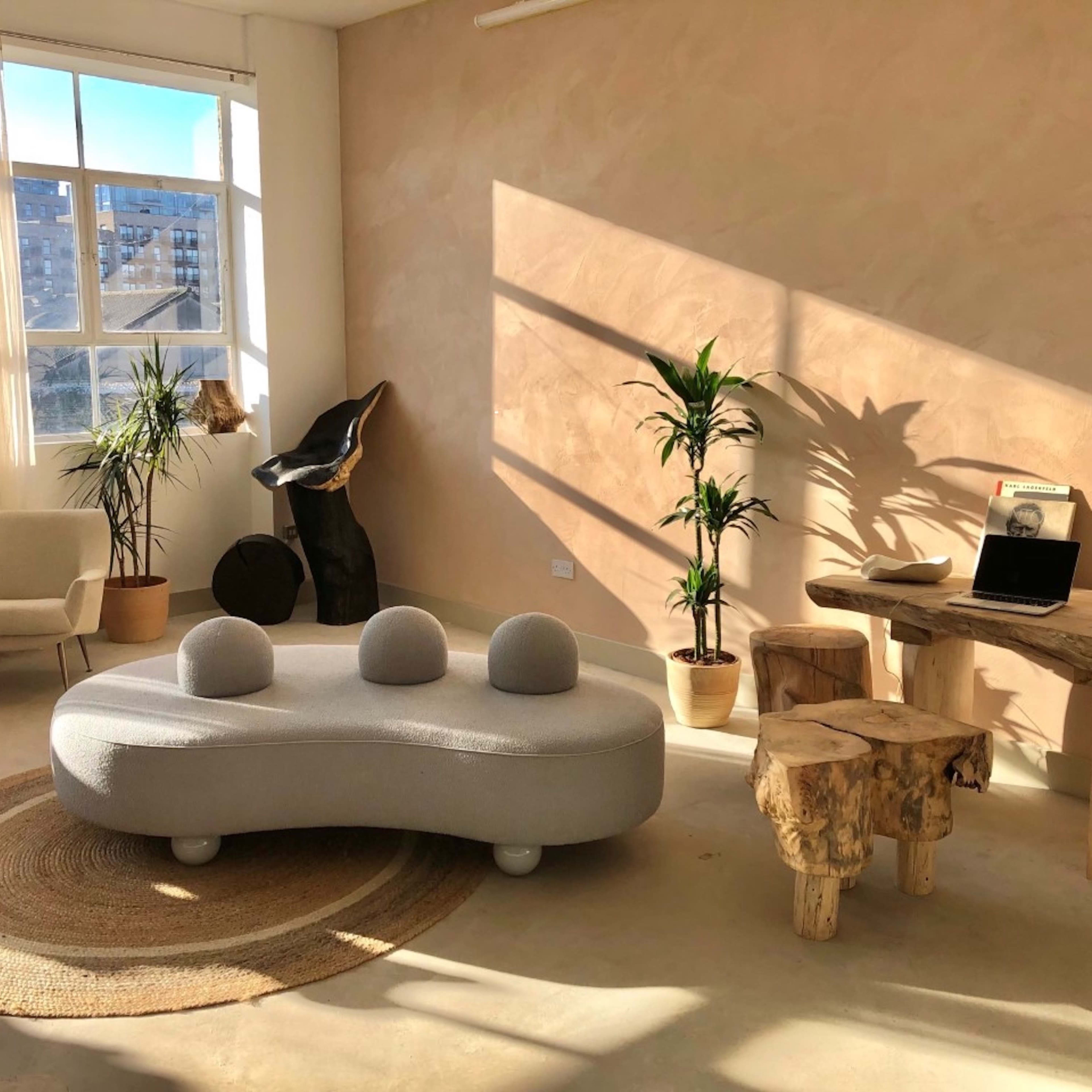 A modern, minimalistic living space with a light-colored wall, a circular gray sofa, natural wood furniture, and houseplants arranged beside a large window.