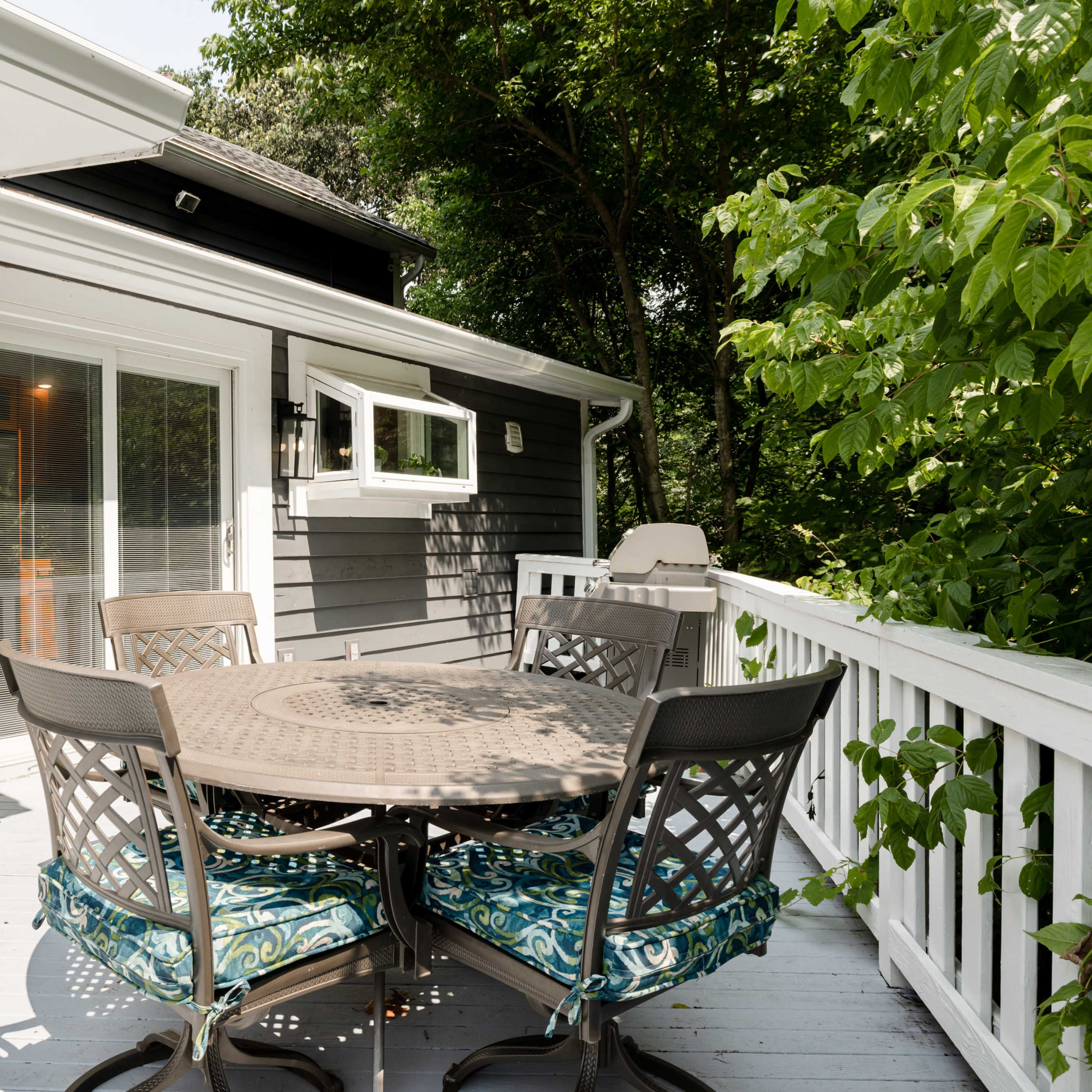 A patio features a round table surrounded by six chairs, set against a backdrop of greenery and a home with gray siding.