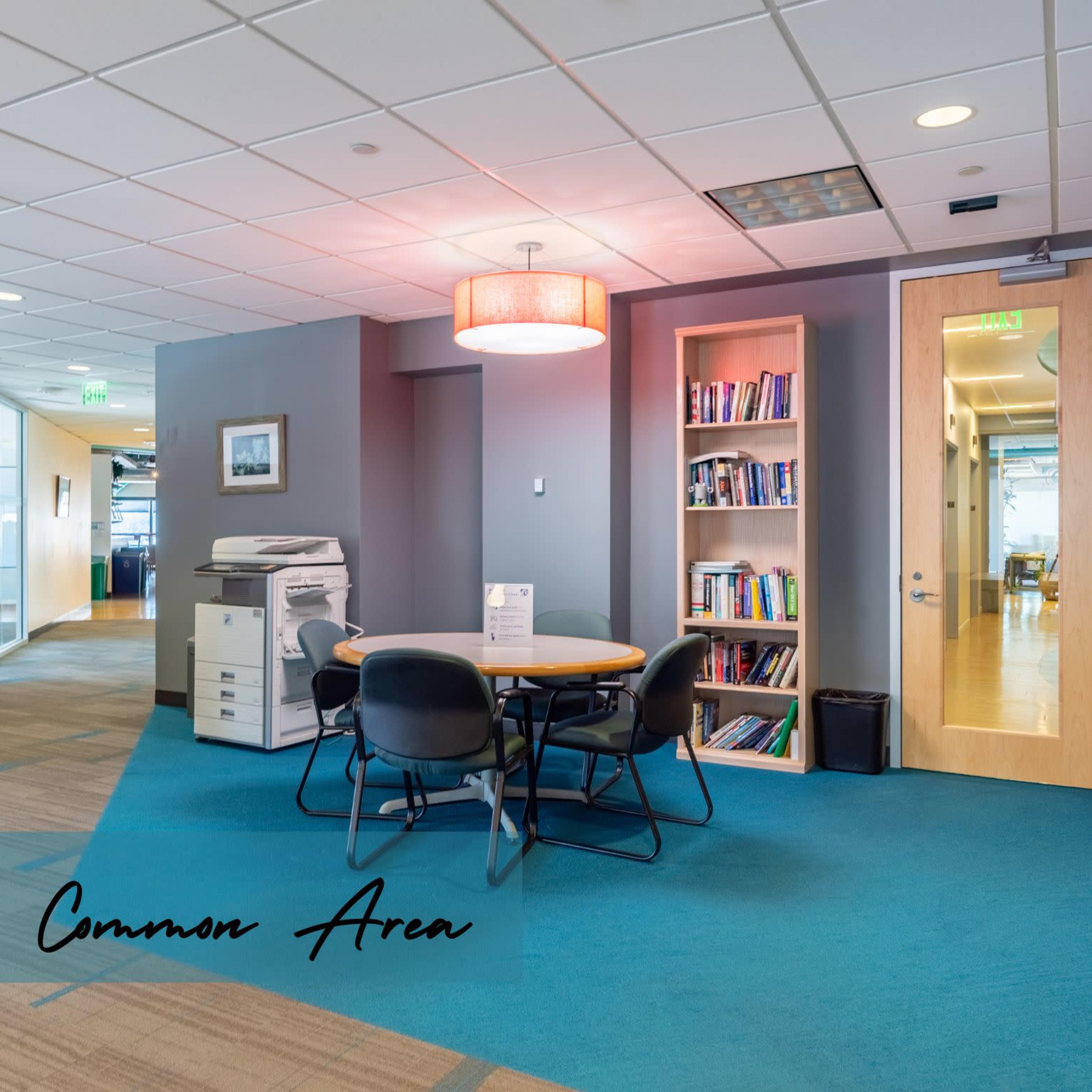 A shared office space with a printer, a round table with chairs, and a bookshelf against a gray wall.