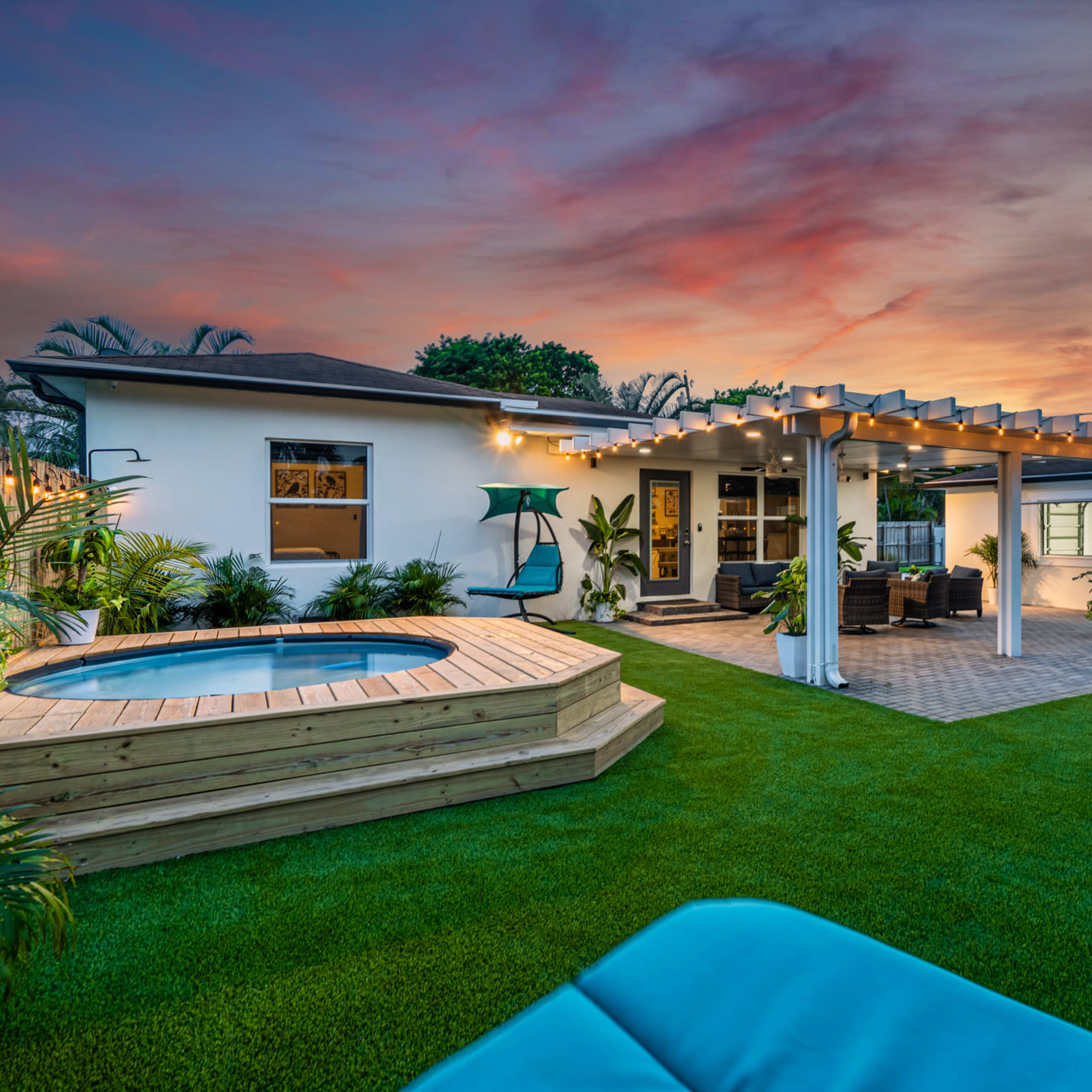 A backyard with a wooden deck, a hot tub, and a spacious lawn, framed by a house with a pergola and patio furniture under a colorful sunset sky.
