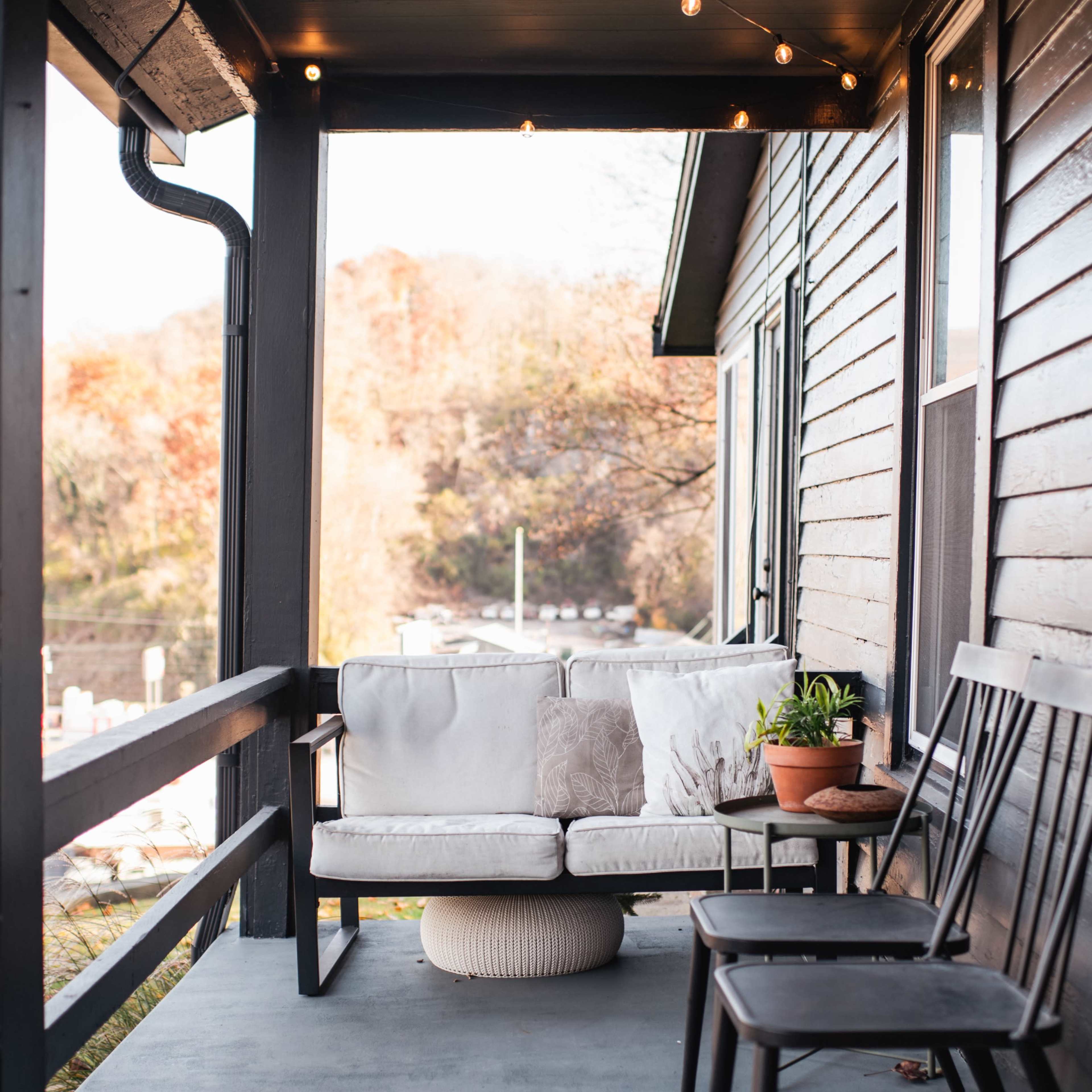 A porch with a white couch, a round ottoman, a potted plant, and a black wooden chair, all under warm string lights.
