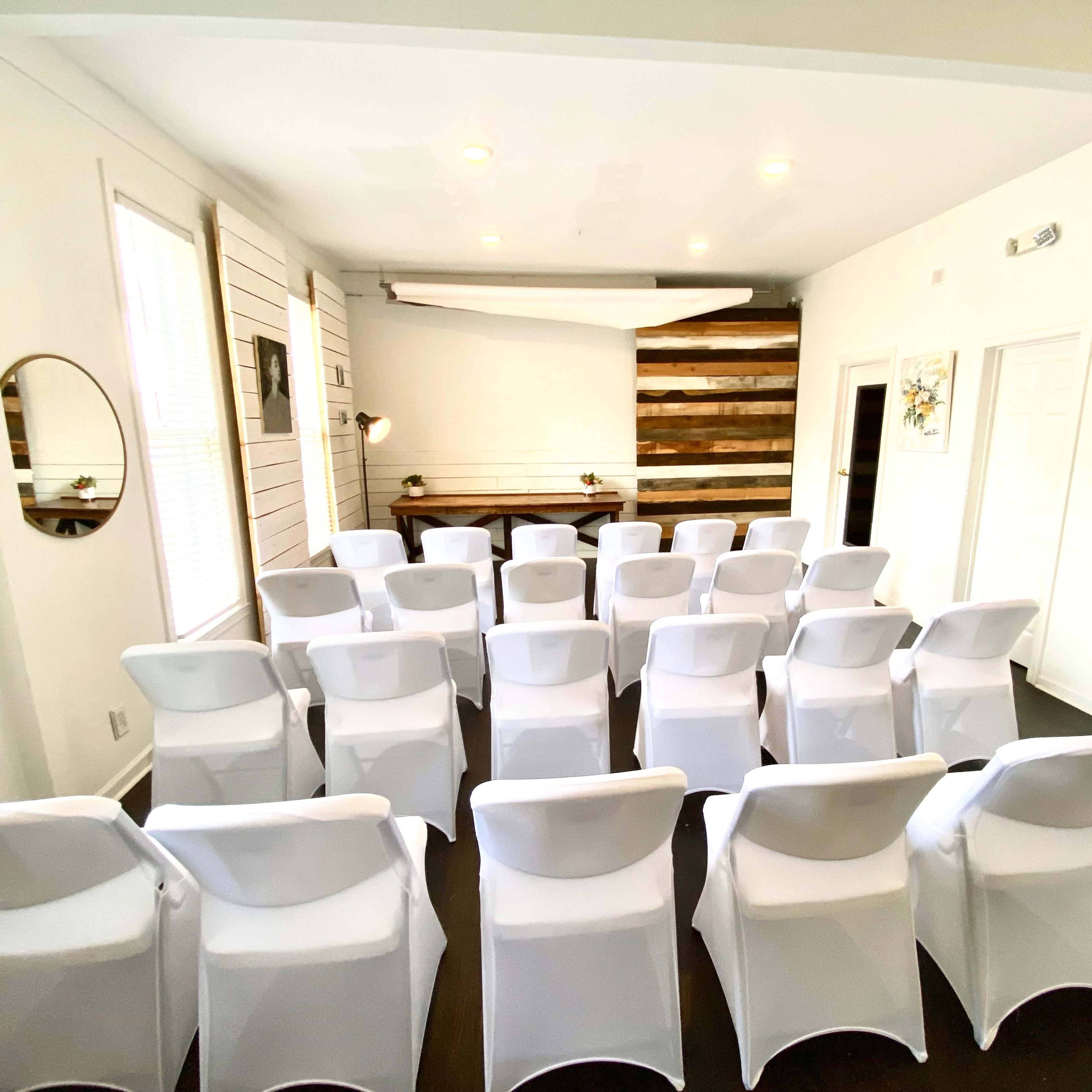 A row of white chairs is set up facing a wooden-paneled wall in a well-lit room.