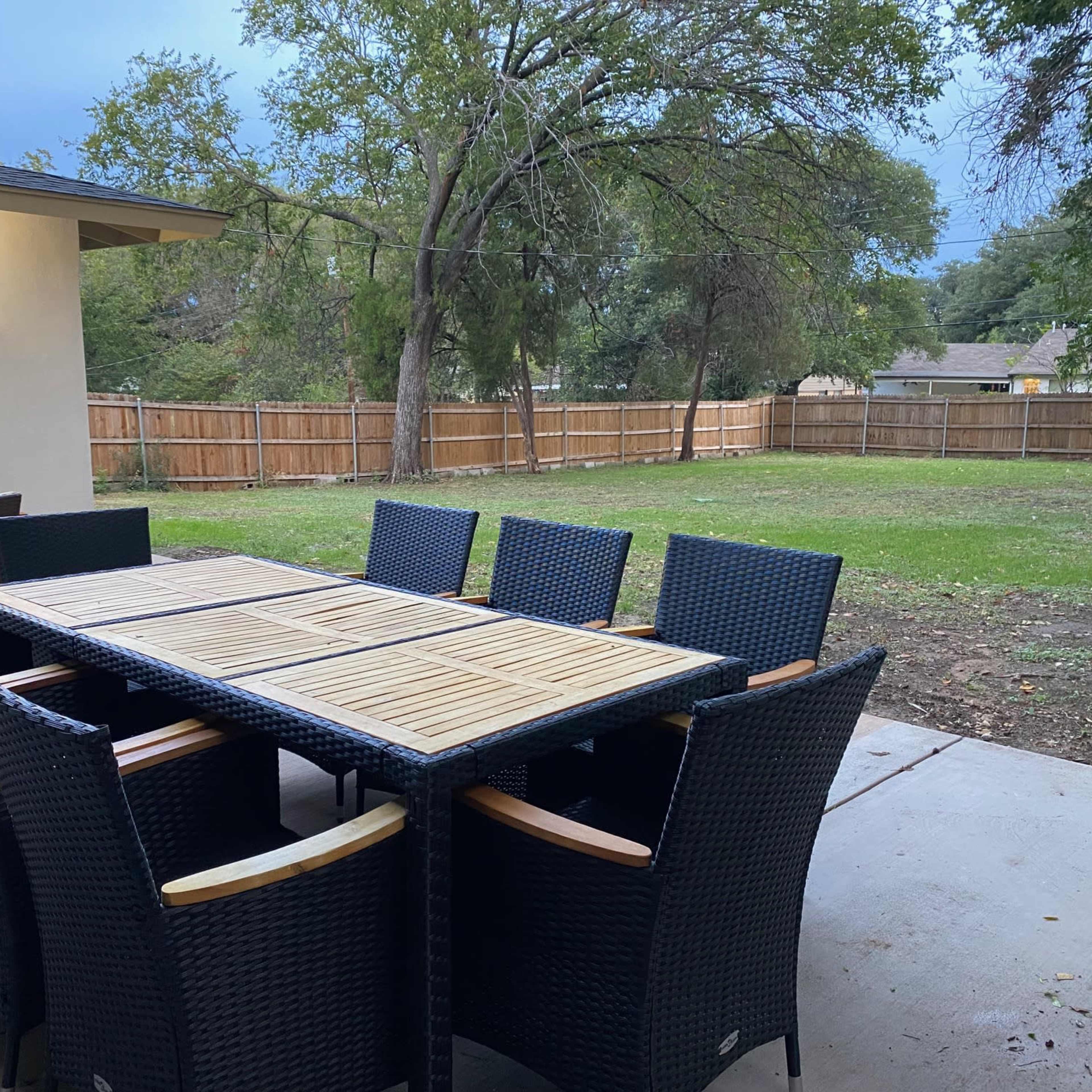 A large outdoor dining table with eight chairs is set on a concrete patio surrounded by a grassy area and a wooden fence.