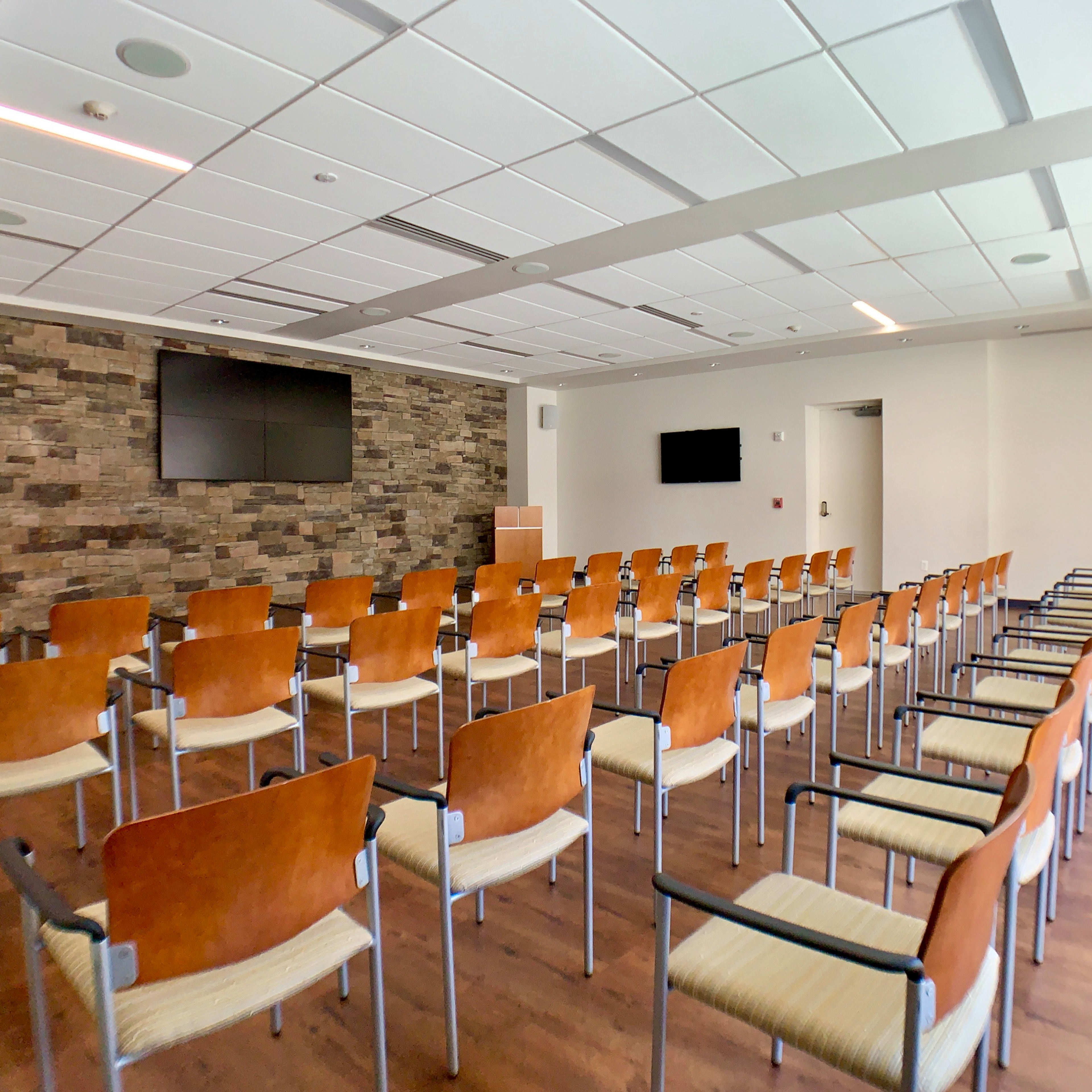 A conference room with rows of chairs arranged facing two large screens on a stone wall.