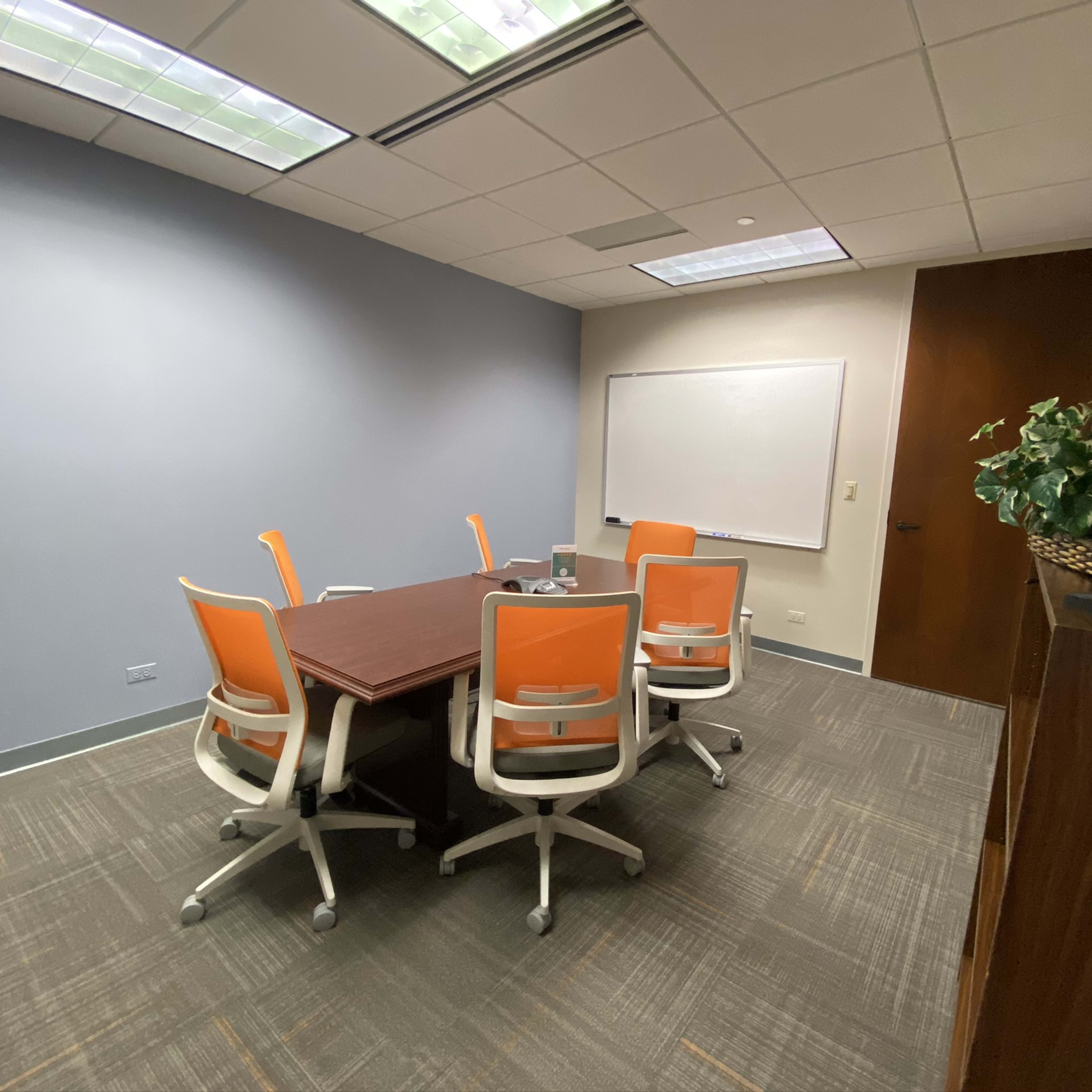 A conference room features a rectangular table surrounded by orange and white chairs, with a whiteboard on one wall and a plant in a pot.