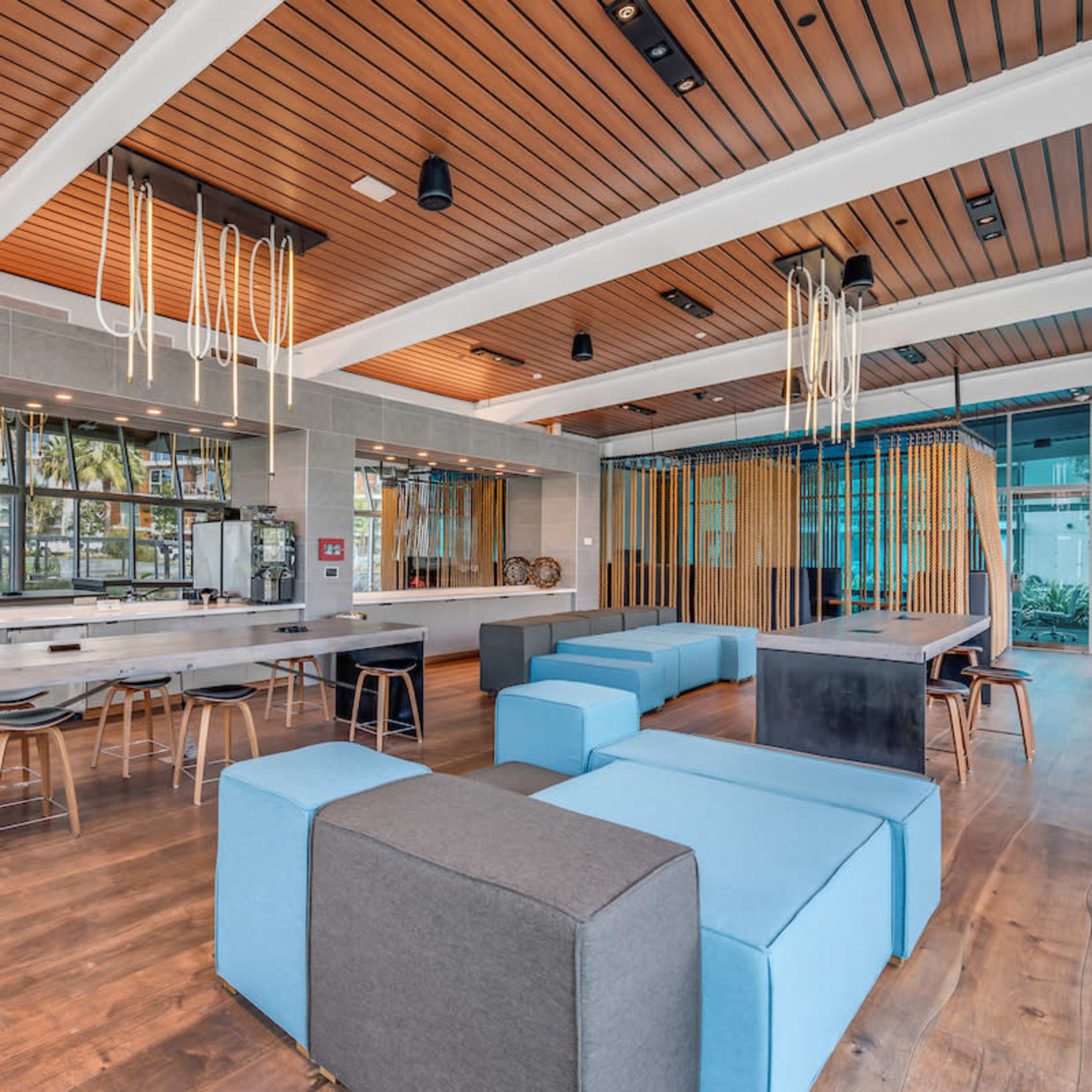 A modern, open-concept communal space with wooden flooring, large windows, a bar area with high stools, and various seating cubes in blue and gray.