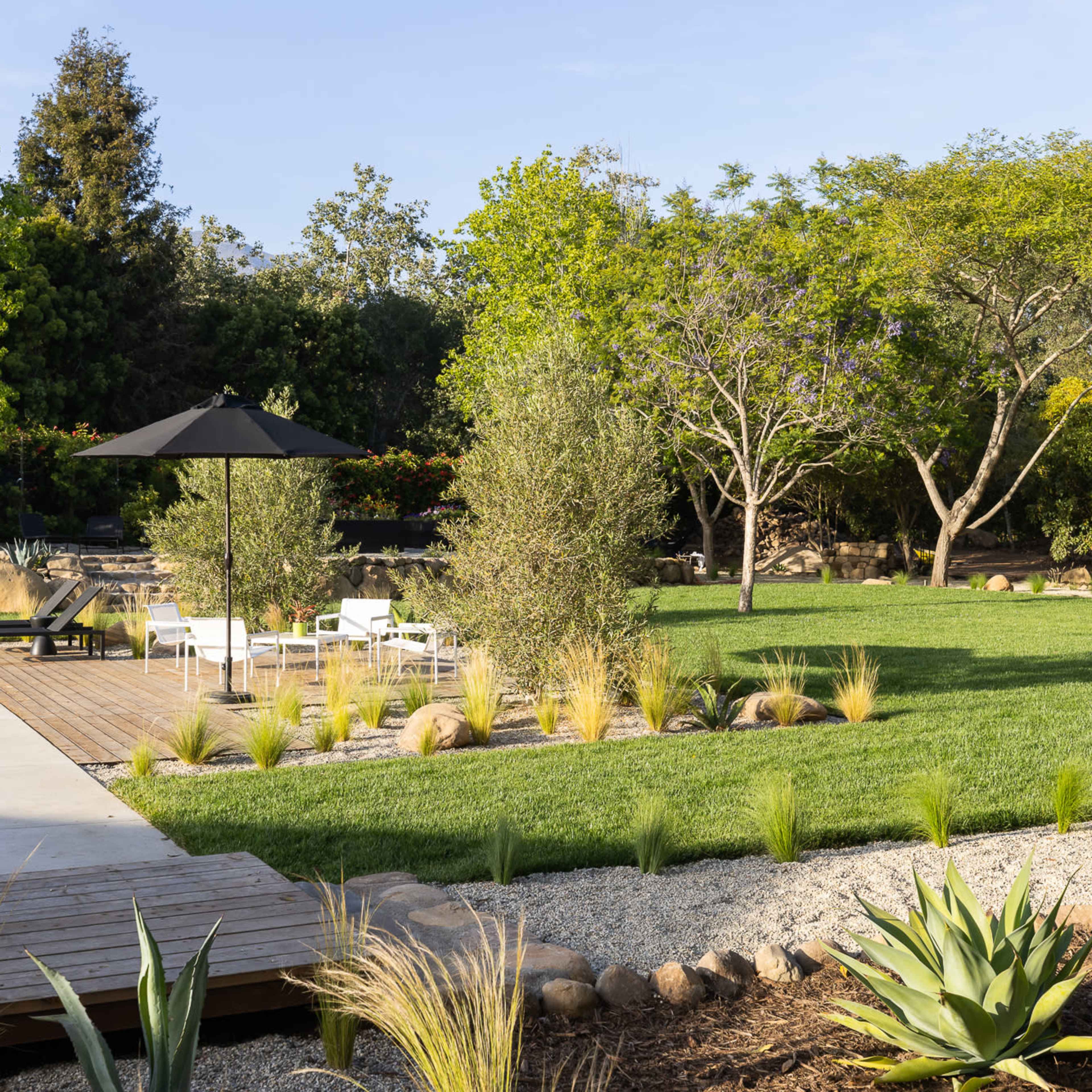 A landscaped garden featuring a wooden deck, green lawn, and scattered rocks, with lounge chairs and an umbrella.