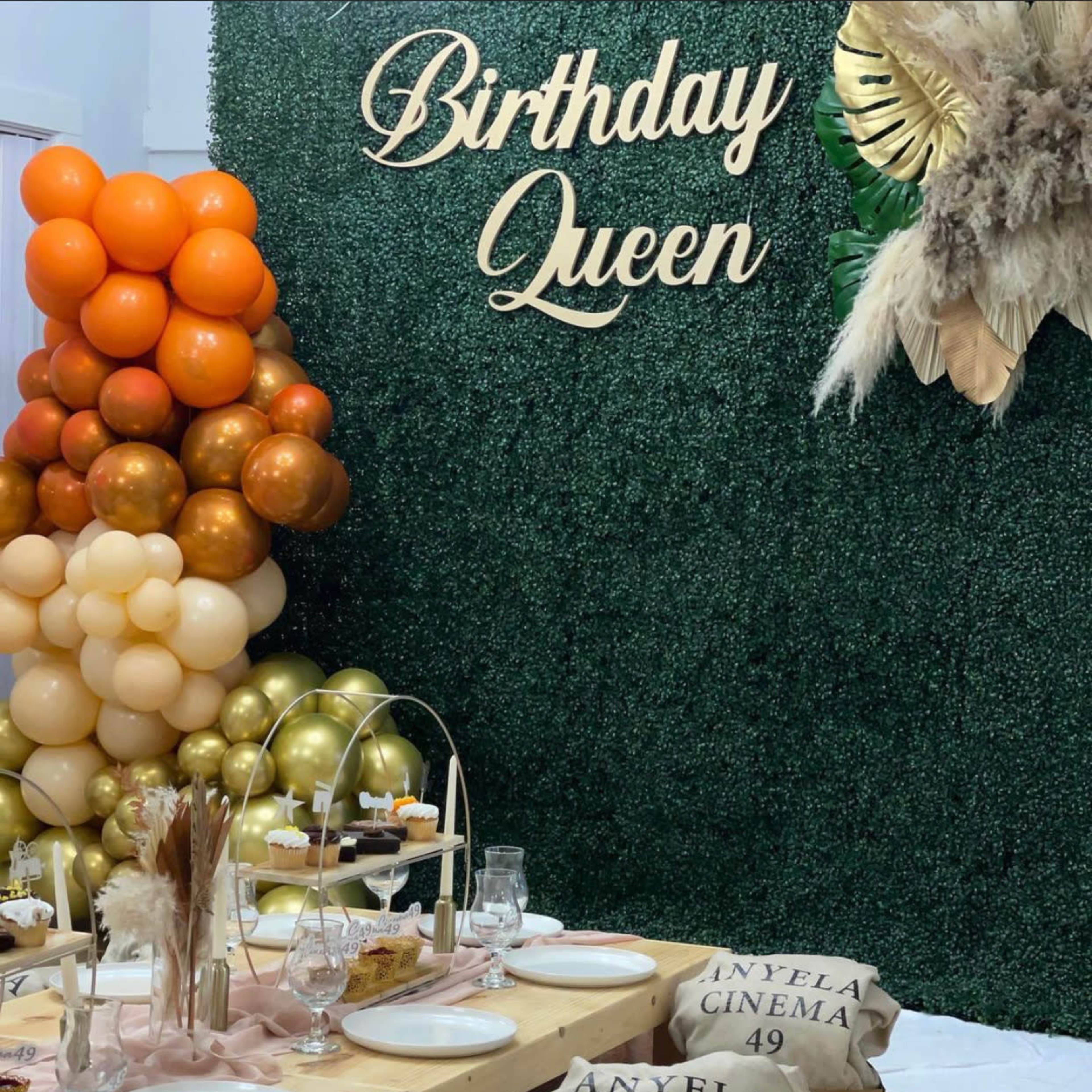 A decorated birthday celebration table features a backdrop of greenery and a balloon arch, with personalized place settings and a "Birthday Queen" sign.