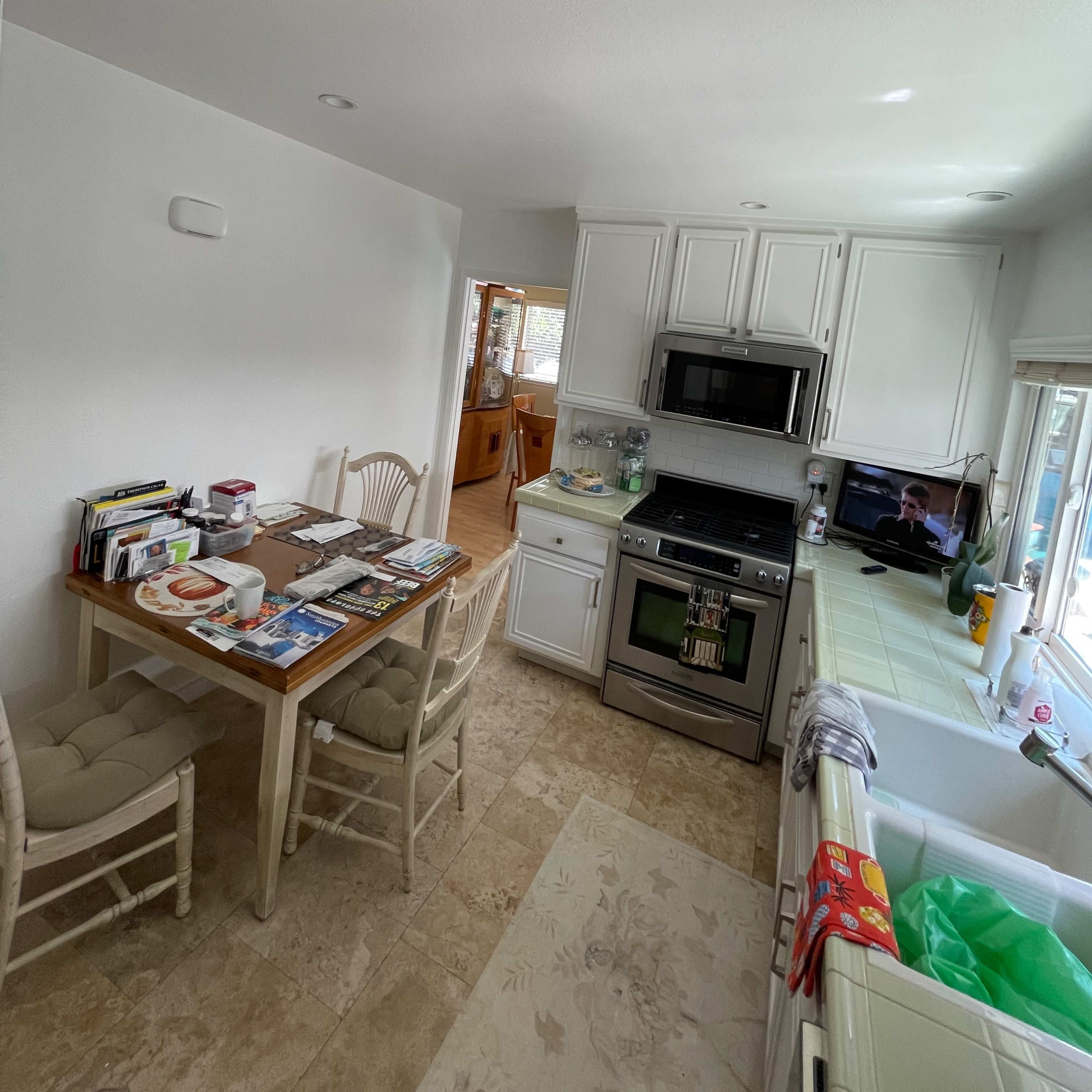 A kitchen with white cabinetry and a wooden dining table surrounded by chairs, featuring various items on the table and an oven in the corner.