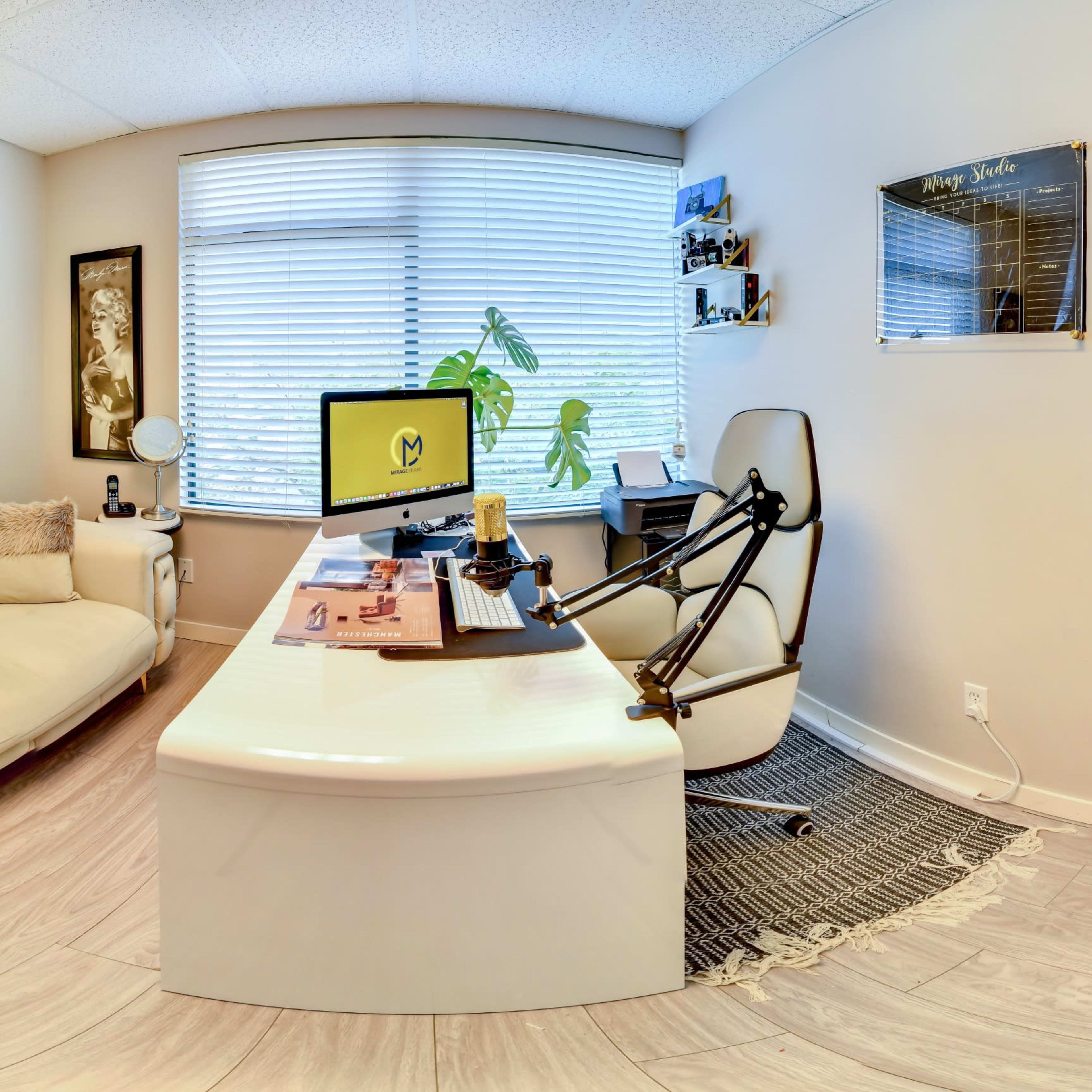 A modern office space featuring a light-colored couch, a large window with a plant, and a desk equipped with a computer and microphone setup, surrounded by shelves displaying various items.