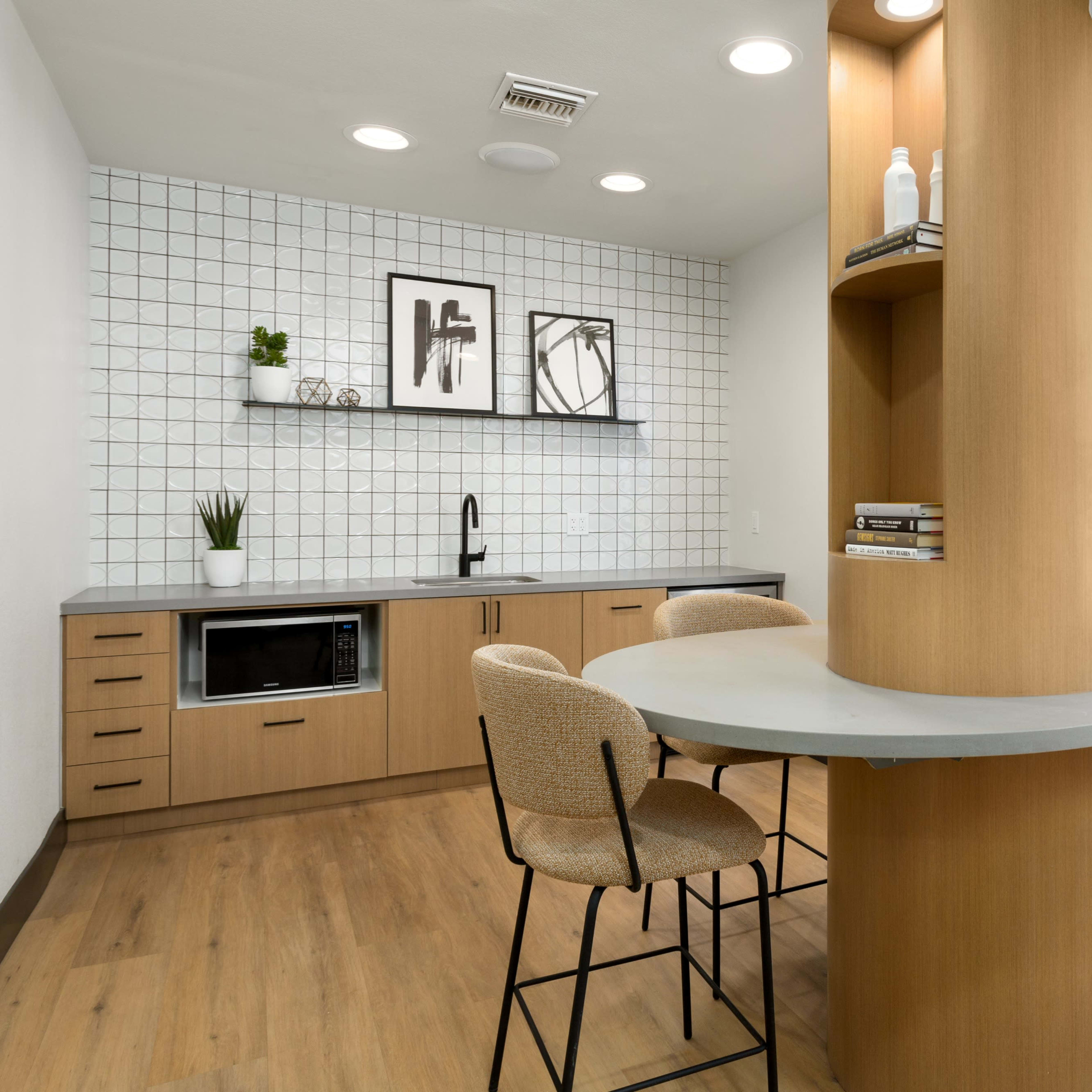 A modern kitchen area with light wood cabinetry, a gray countertop, a microwave, and a small dining table with two bar stools.