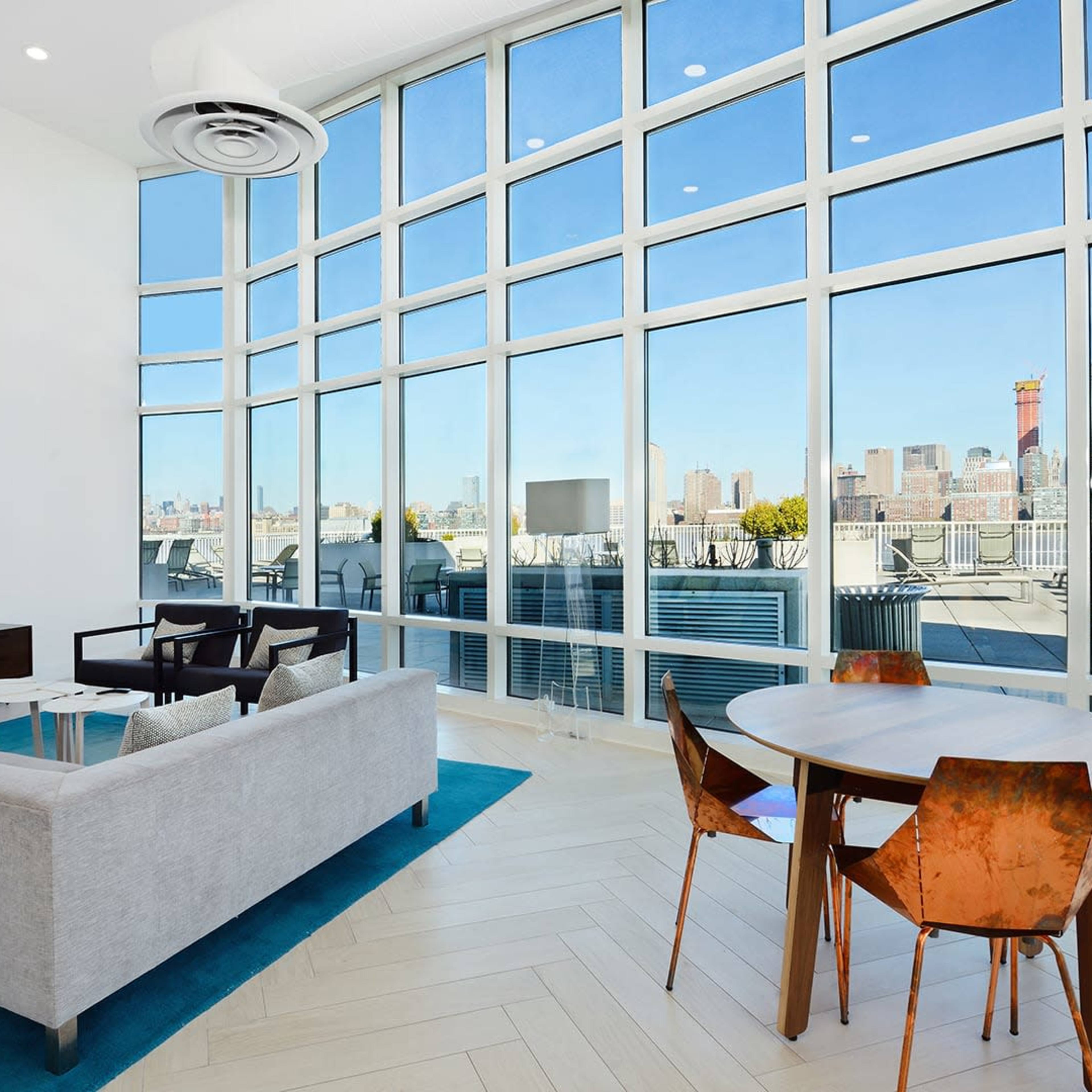 The modern living space features a sectional sofa, a small wooden dining table, and large windows with a view of a city skyline.