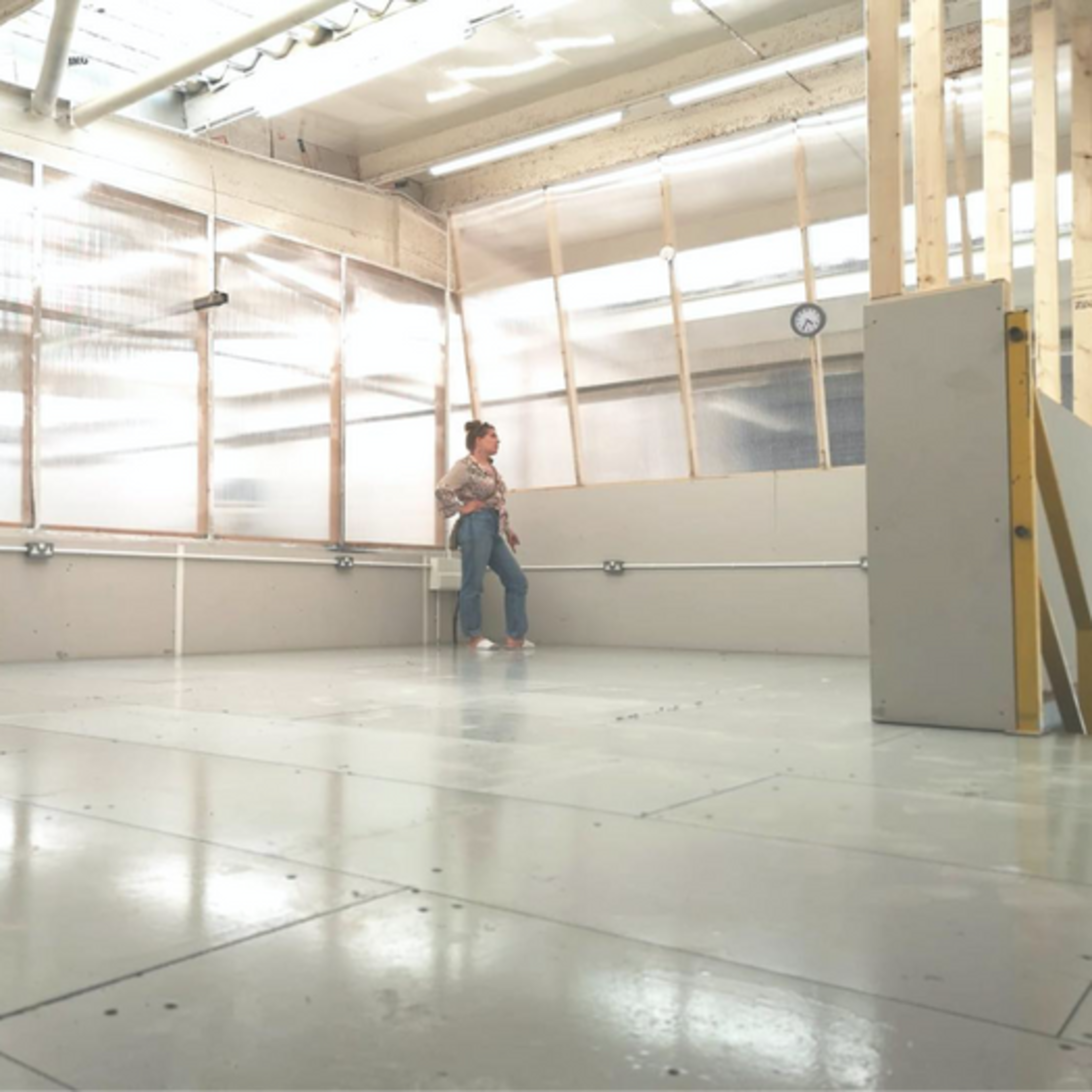 A person stands against a wall in a spacious, unfinished room with large windows and a concrete floor.