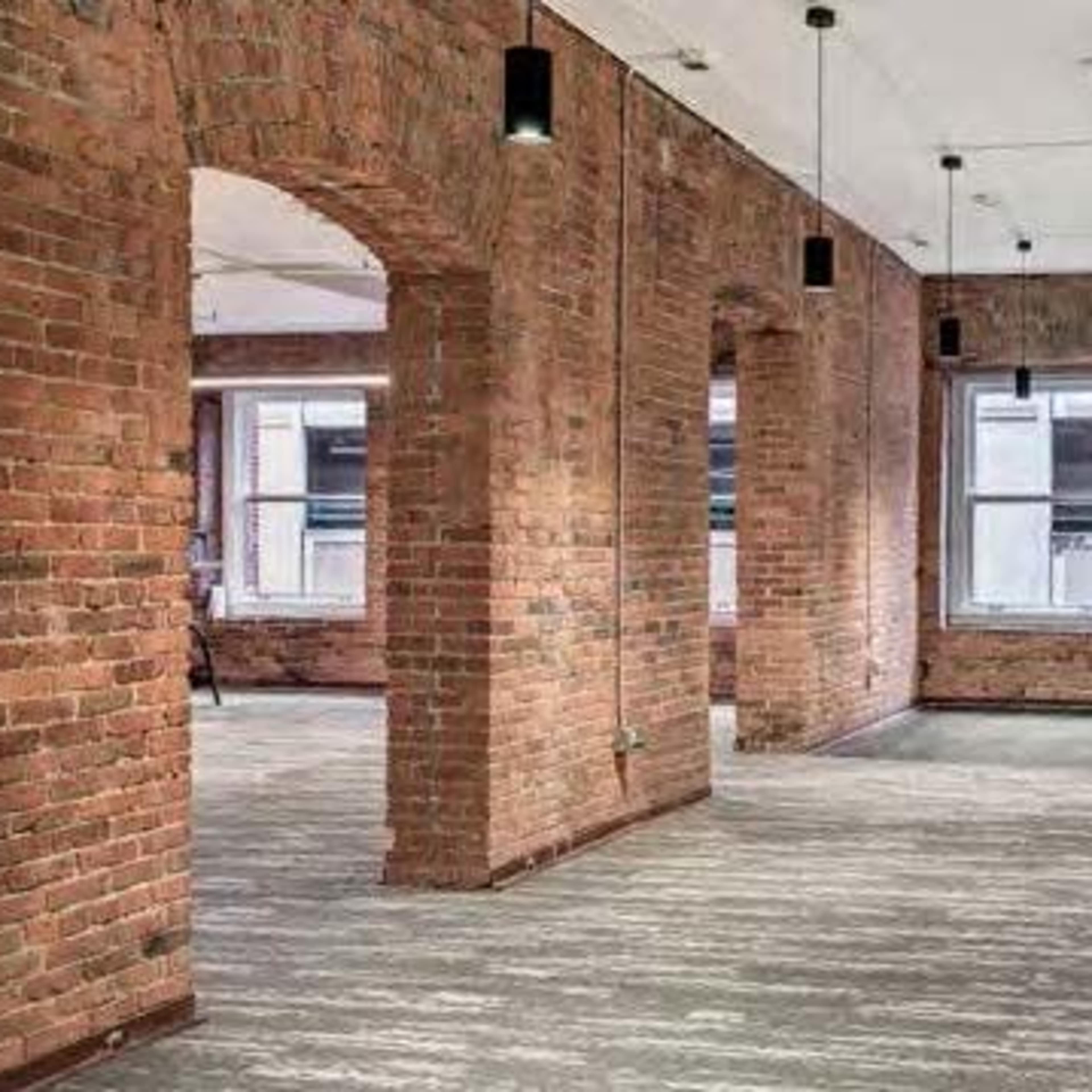 An empty office space featuring exposed brick walls, large windows, and a combination of hardwood and carpet flooring.