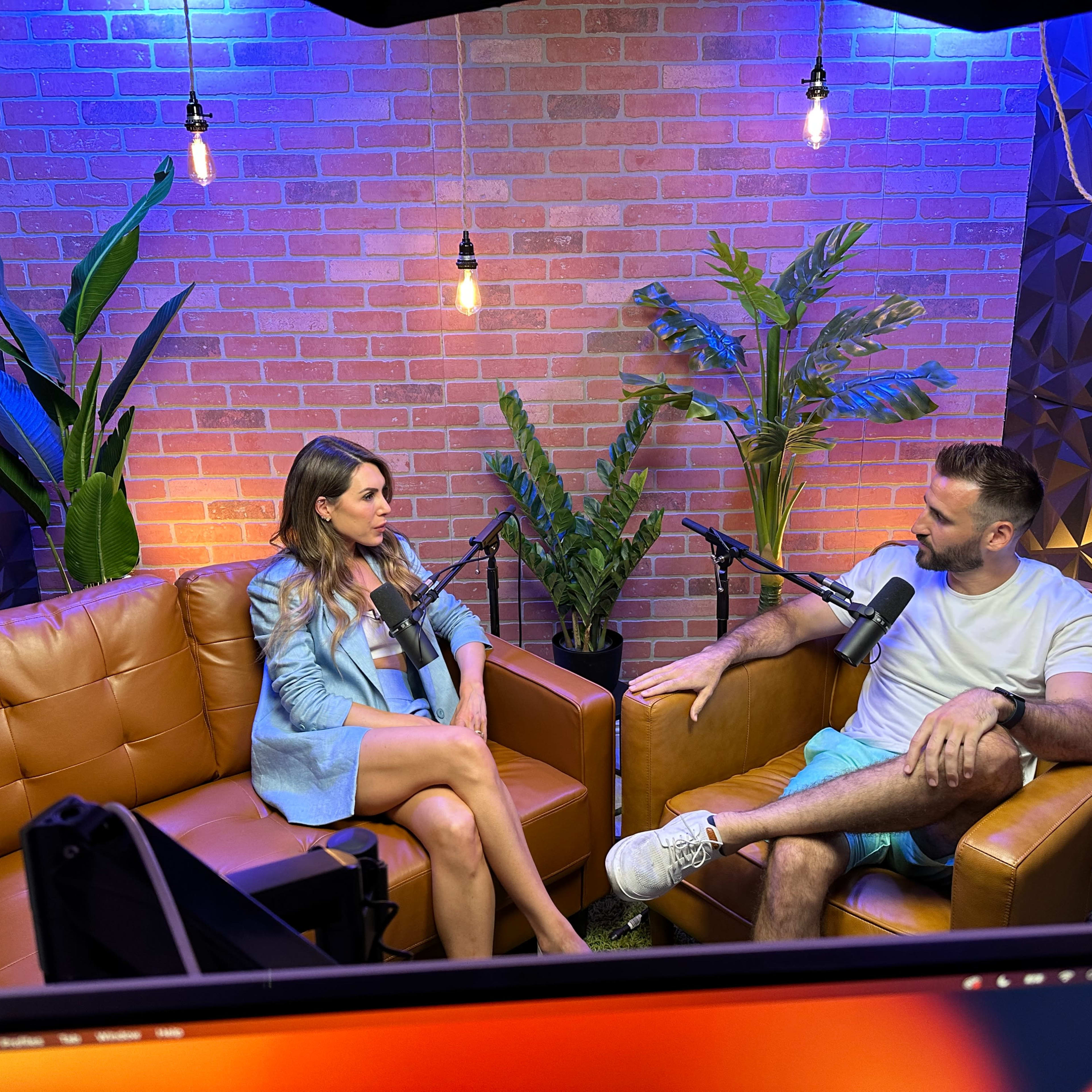 A woman and a man sit on brown couches in a well-lit studio, engaged in conversation with microphones in front of them, surrounded by plants and decorative brick walls.