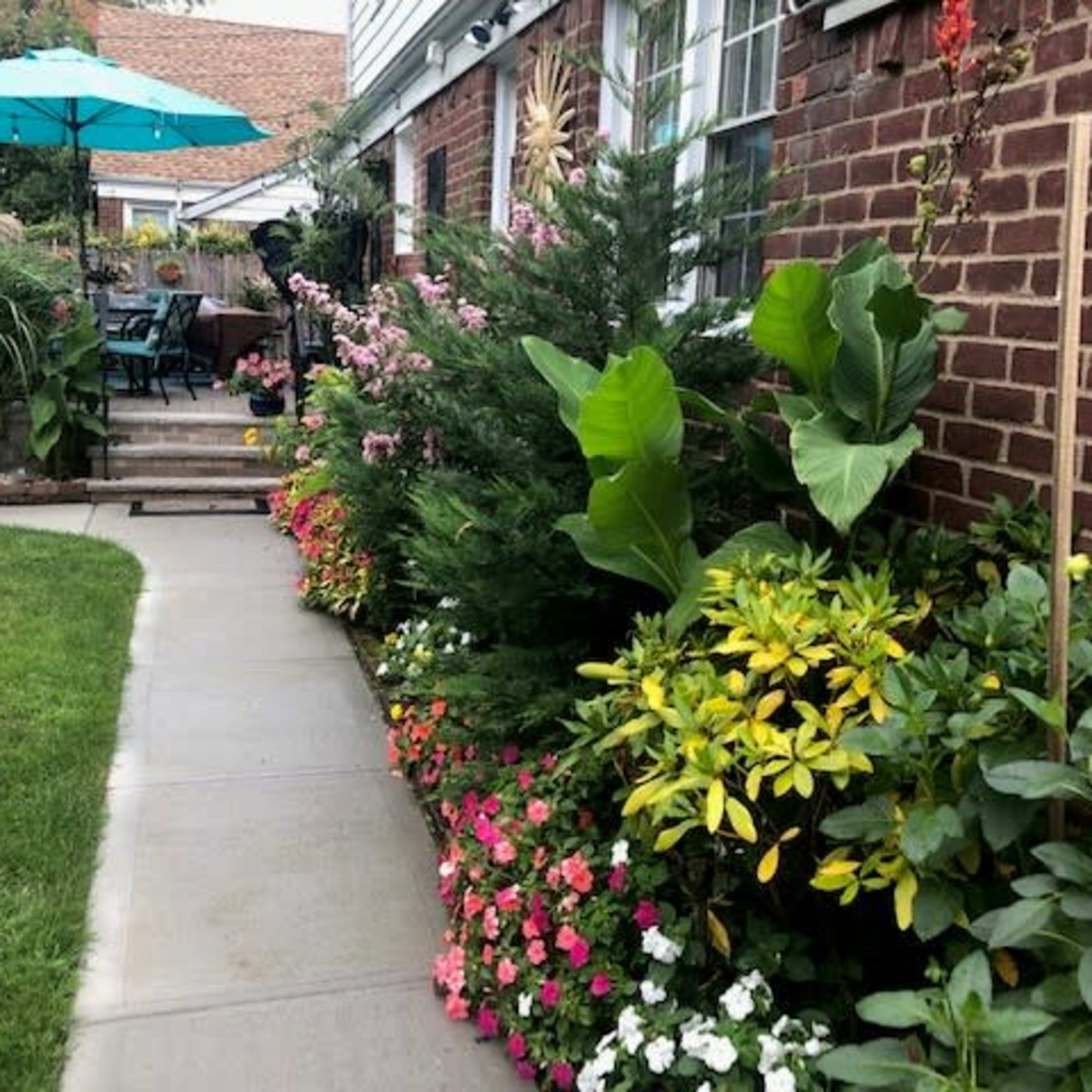 A landscaped garden features a stone pathway bordered by colorful flowers and greenery, leading up to a dining area with an umbrella.