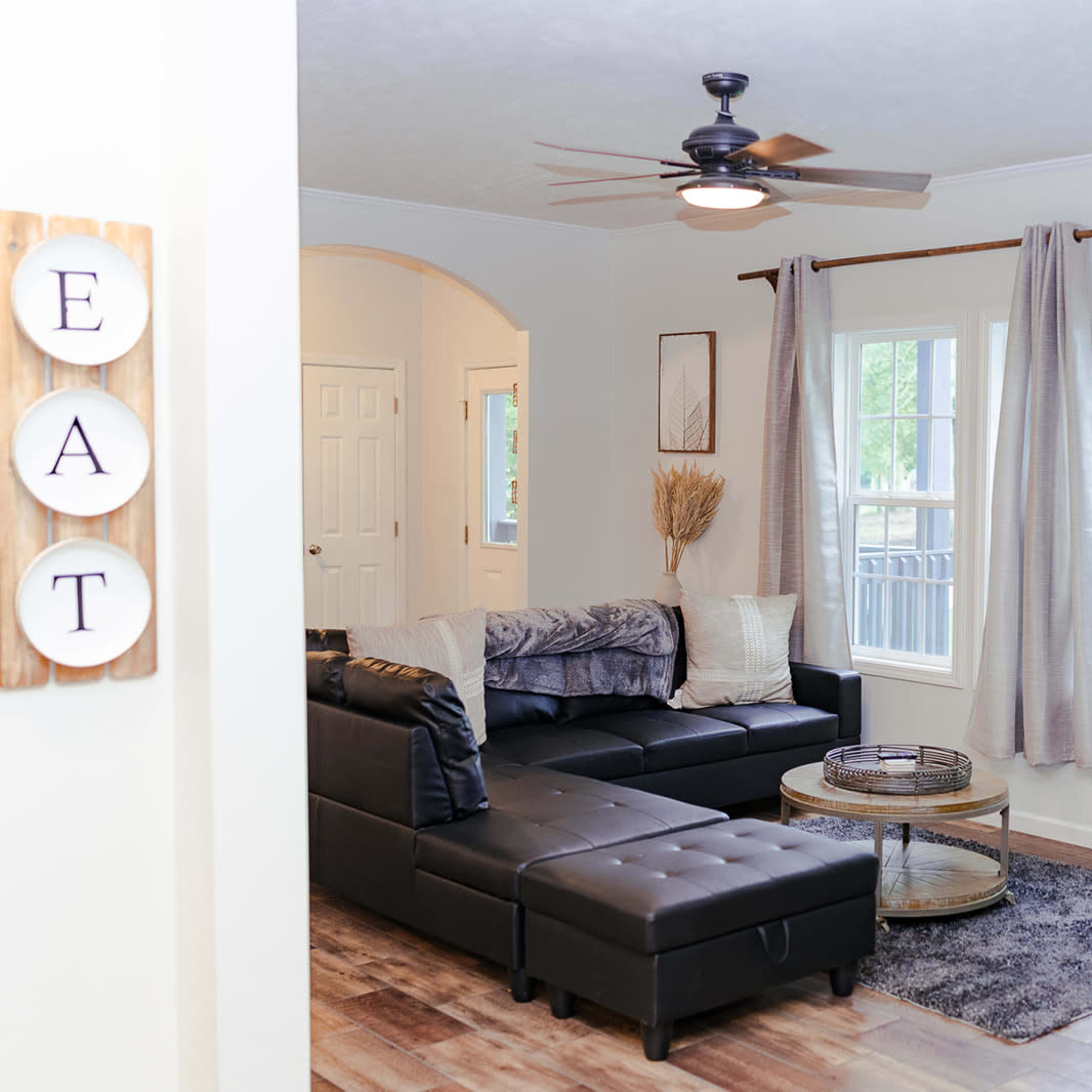 A cozy living room featuring a black sectional sofa, a round coffee table, and large windows with gray curtains.