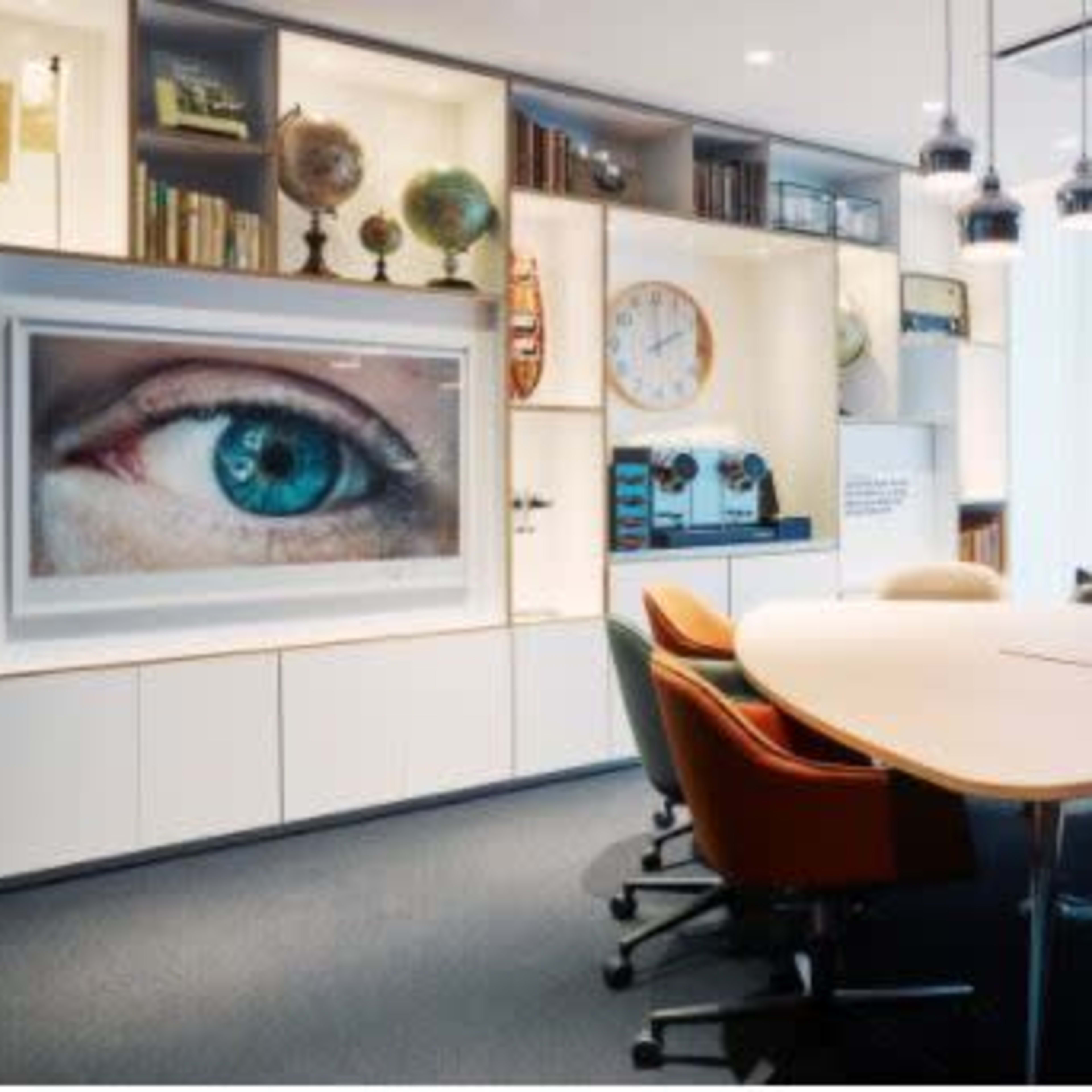 A modern conference room with a large table surrounded by colorful chairs, and a display featuring an eye on the wall among shelves filled with decorative items and a clock.