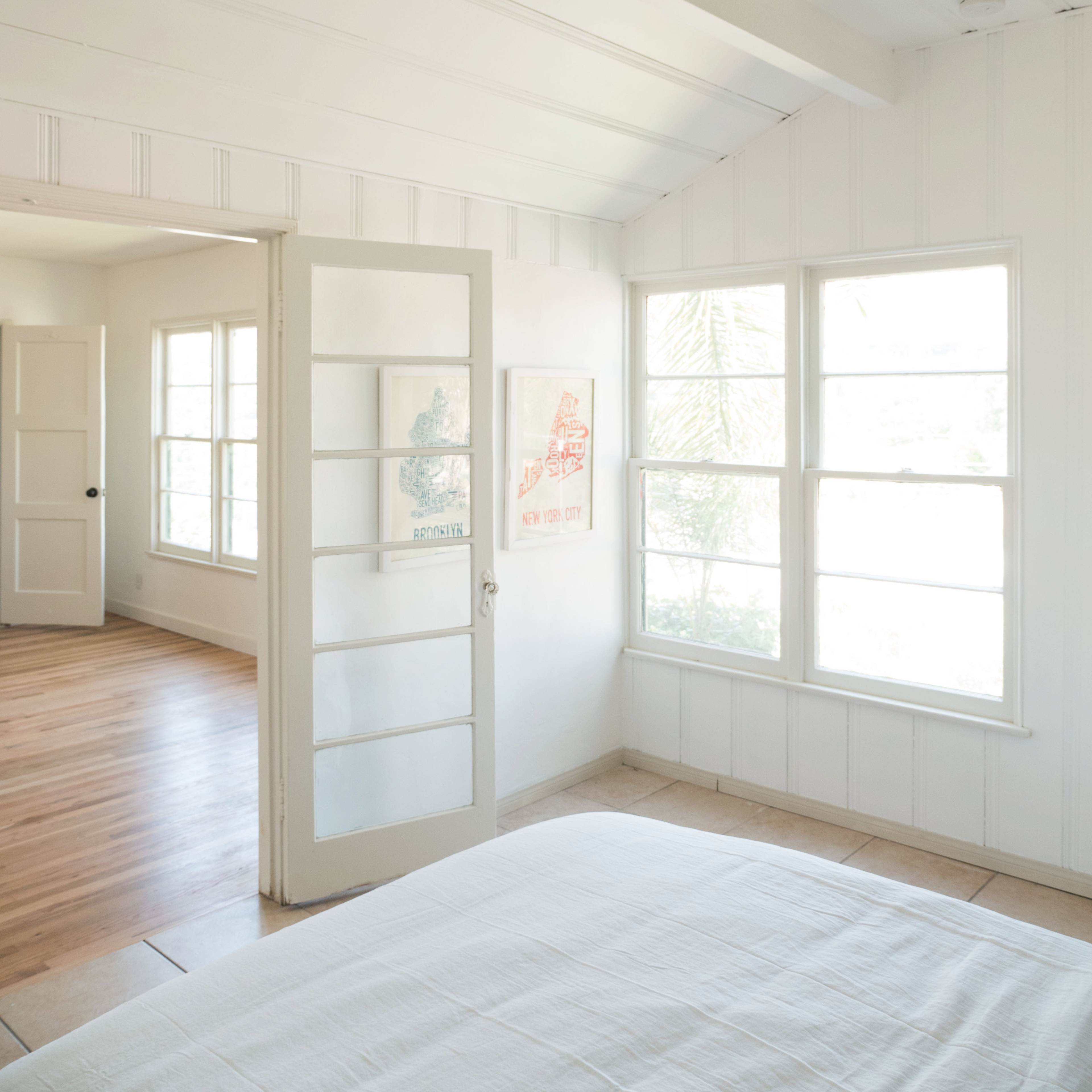 A bright bedroom features large windows and a glass-paneled door leading to another room with wooden flooring.
