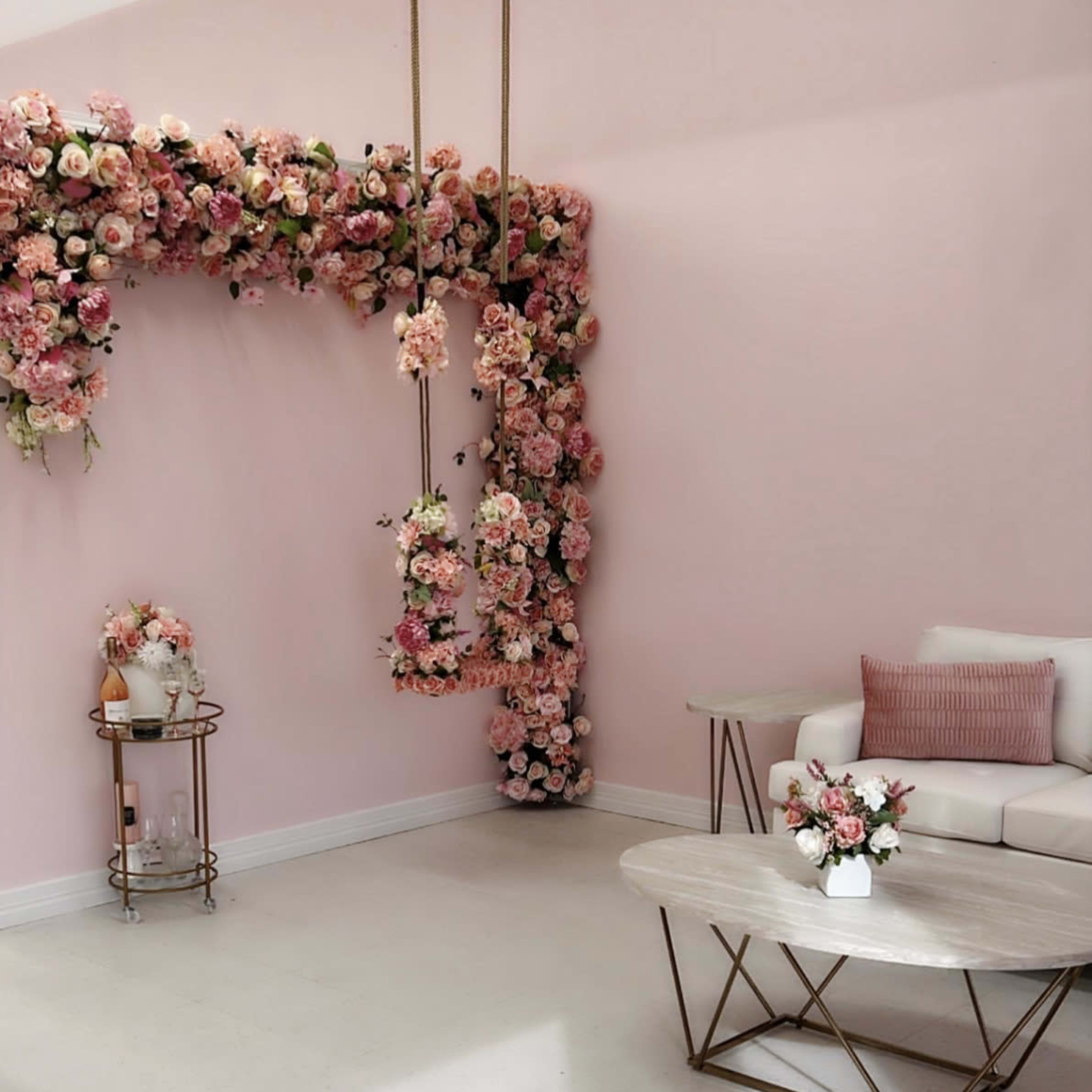 The scene features a light pink room with a floral arch adorned with pink flowers, a white sofa, and a circular coffee table.
