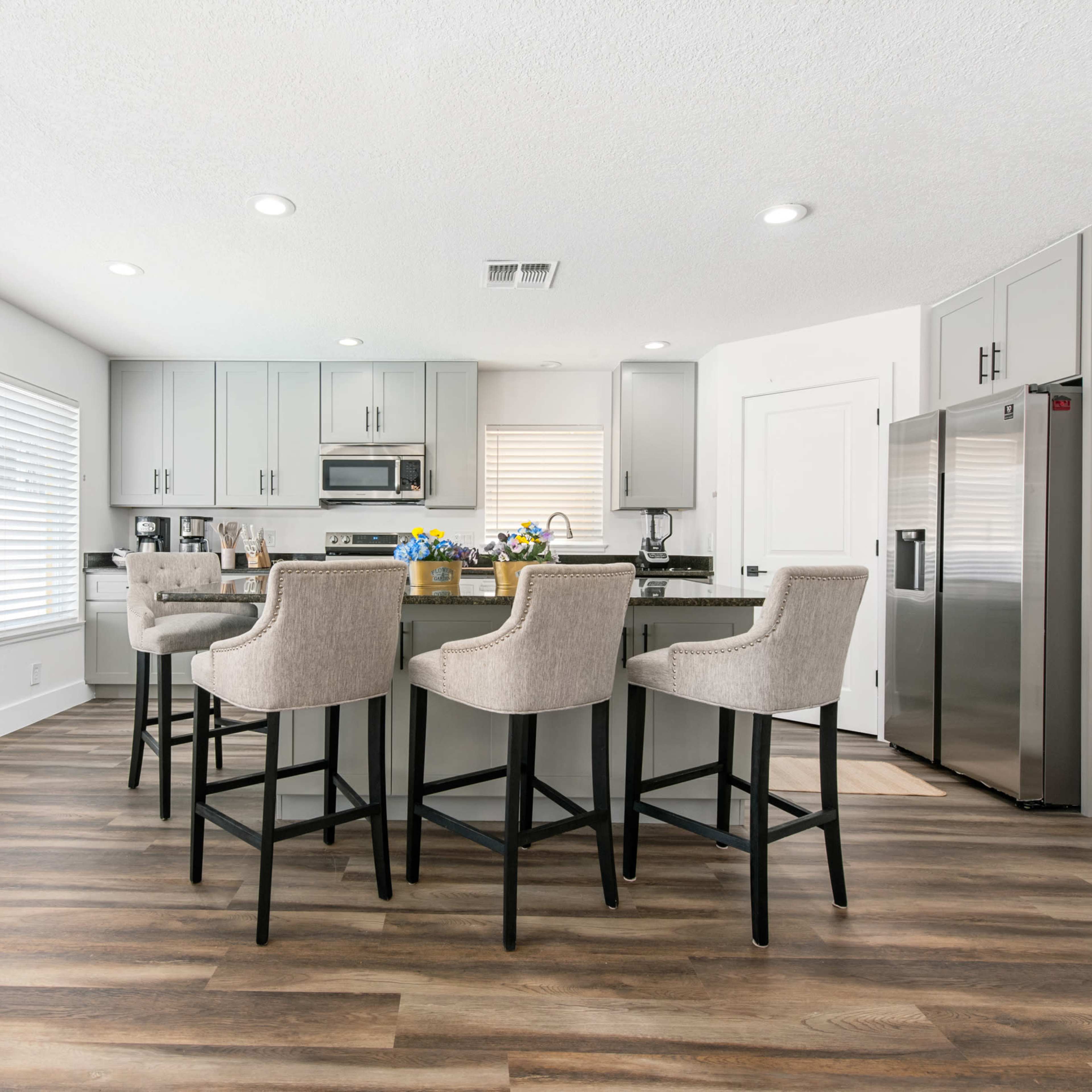 A modern kitchen with gray cabinetry, a stainless steel refrigerator, and four upholstered bar stools around a central island.