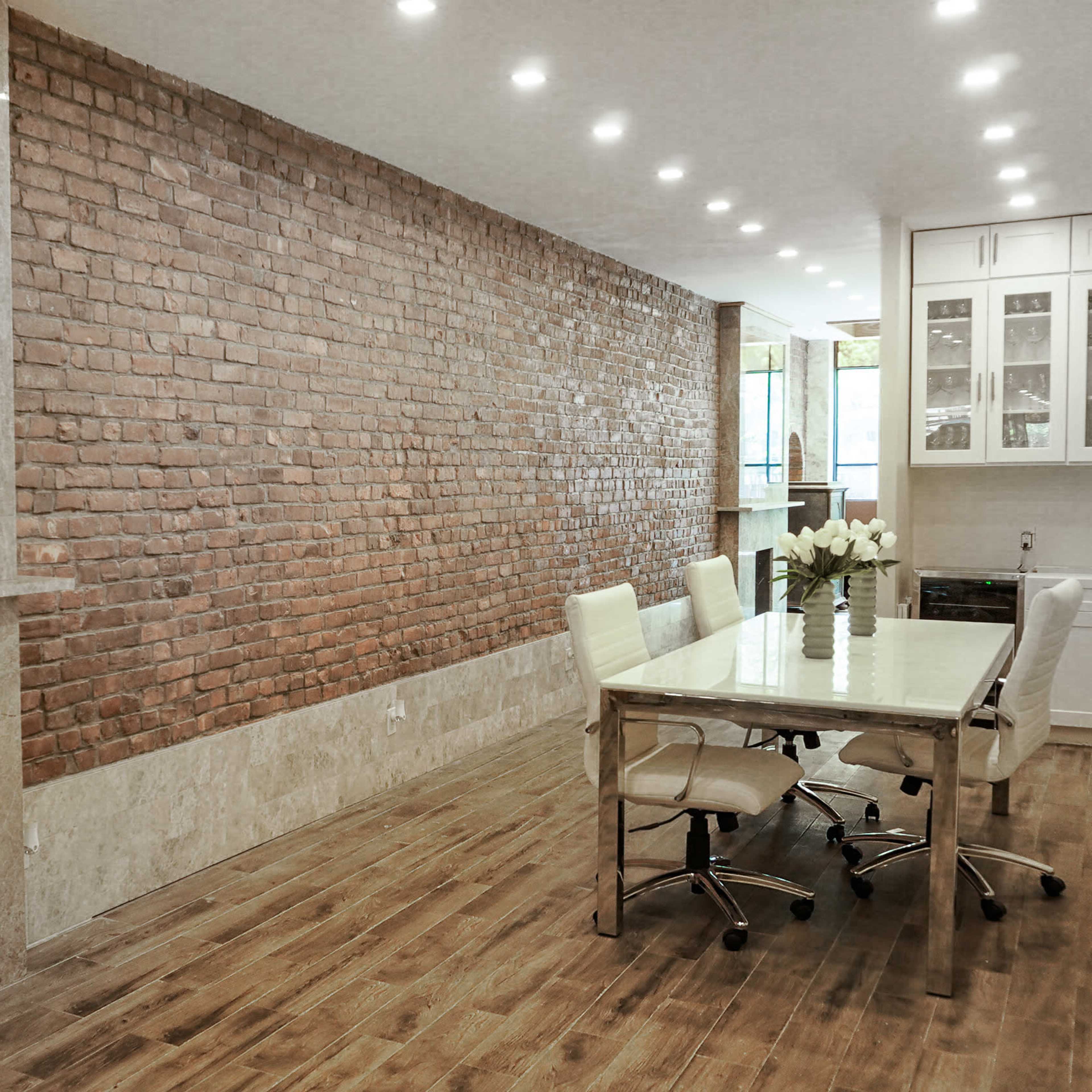 A modern room features a long dining table surrounded by white chairs, with a brick wall and a light-colored marble fireplace.