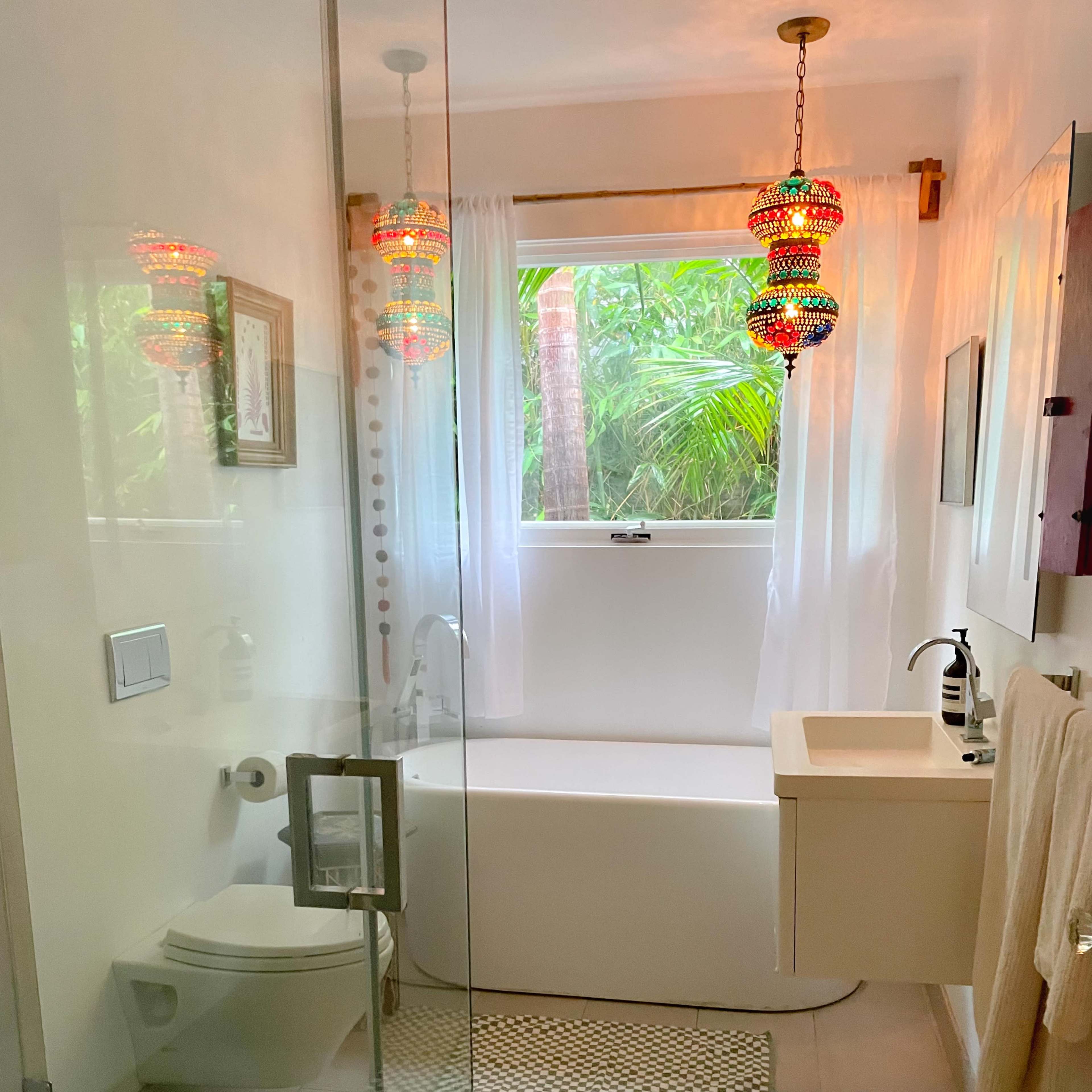 A modern bathroom features a glass shower, a bathtub near a window with greenery outside, and colorful pendant lights.