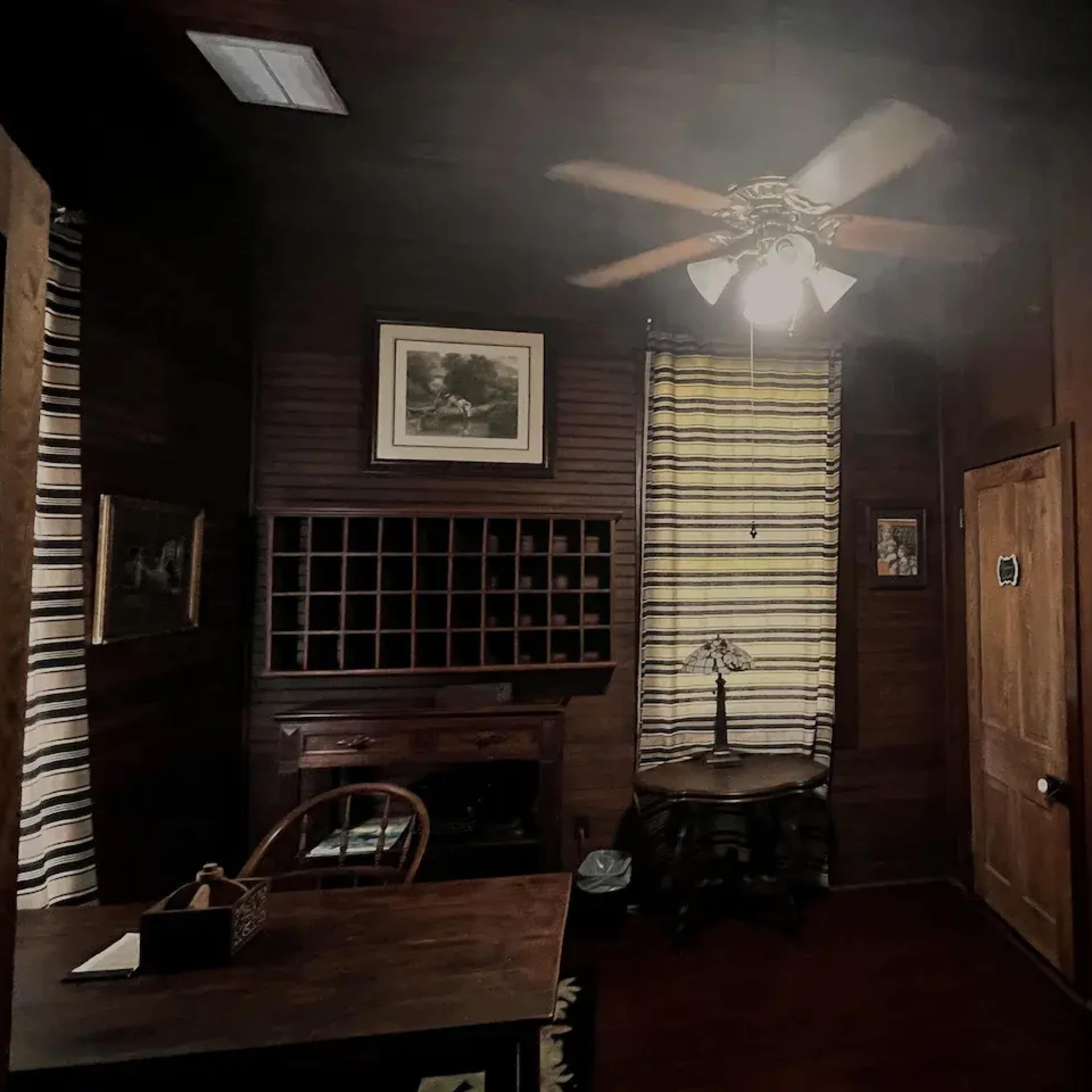 A dimly lit wooden room featuring a ceiling fan, a desk, and two doors, with windows dressed in striped curtains.