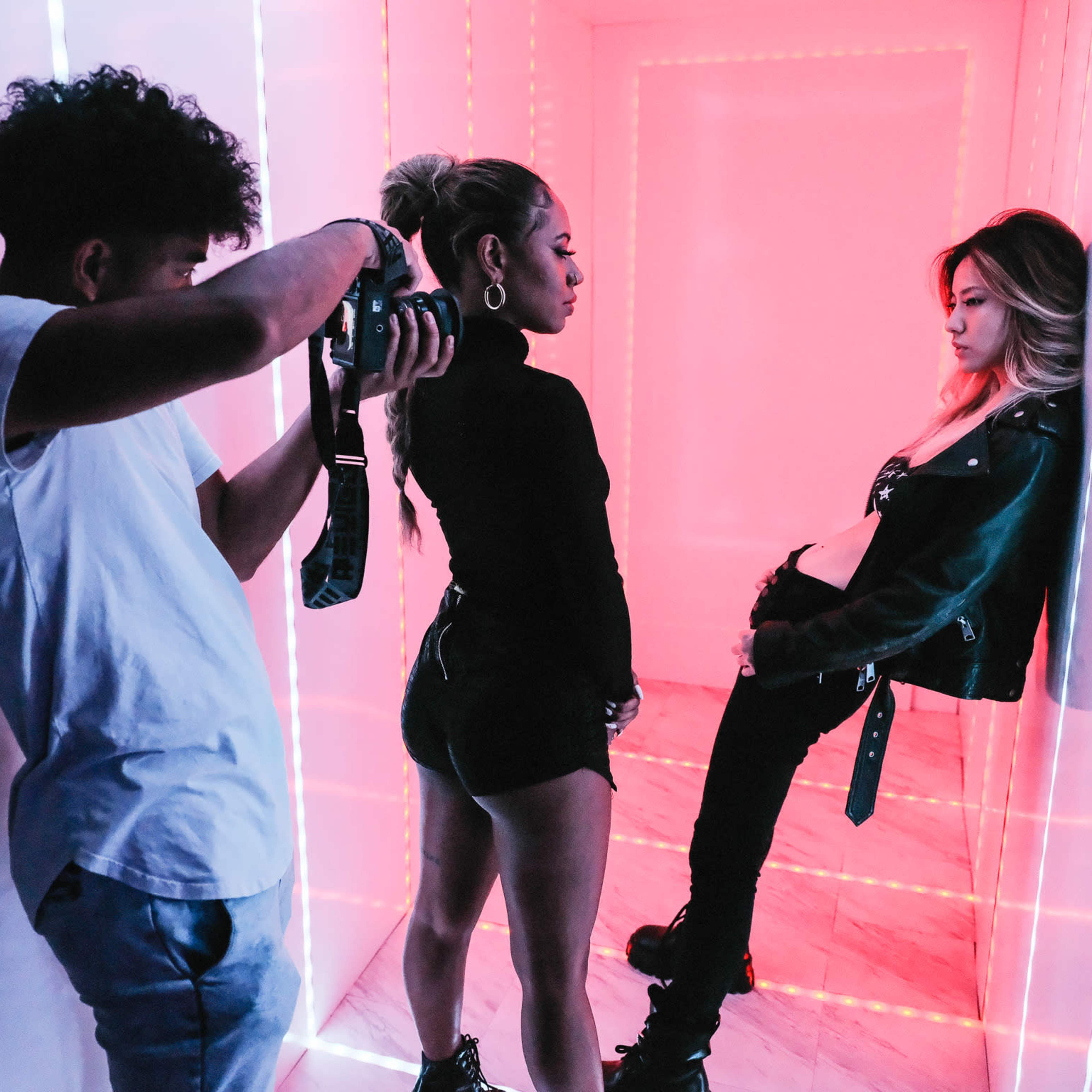 A photographer captures a model posing against a brightly lit, neon pink backdrop while another person stands beside her.