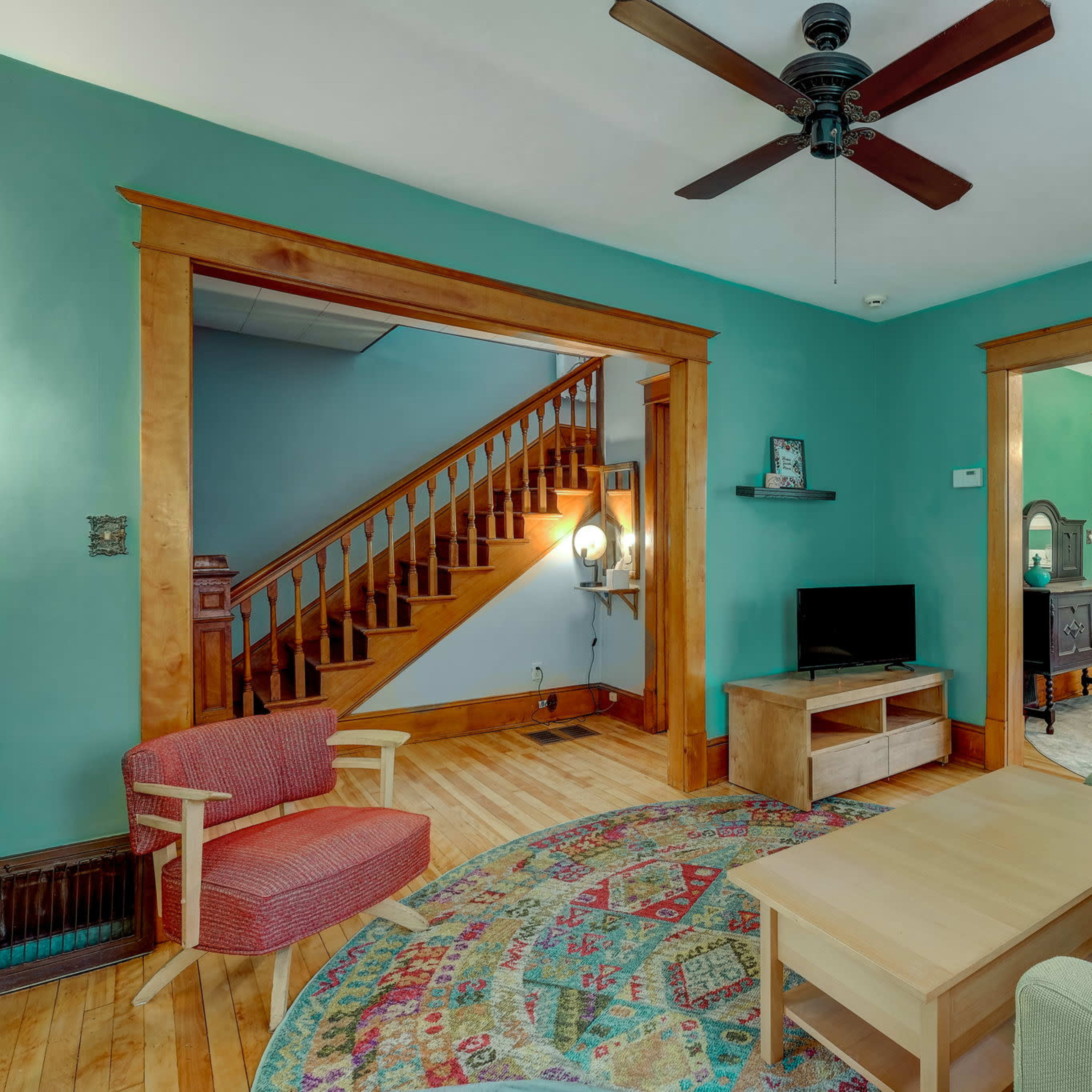 A living room features a teal wall, wooden trim, a ceiling fan, a coffee table, a couch, a chair, and a view of a staircase and dining area.