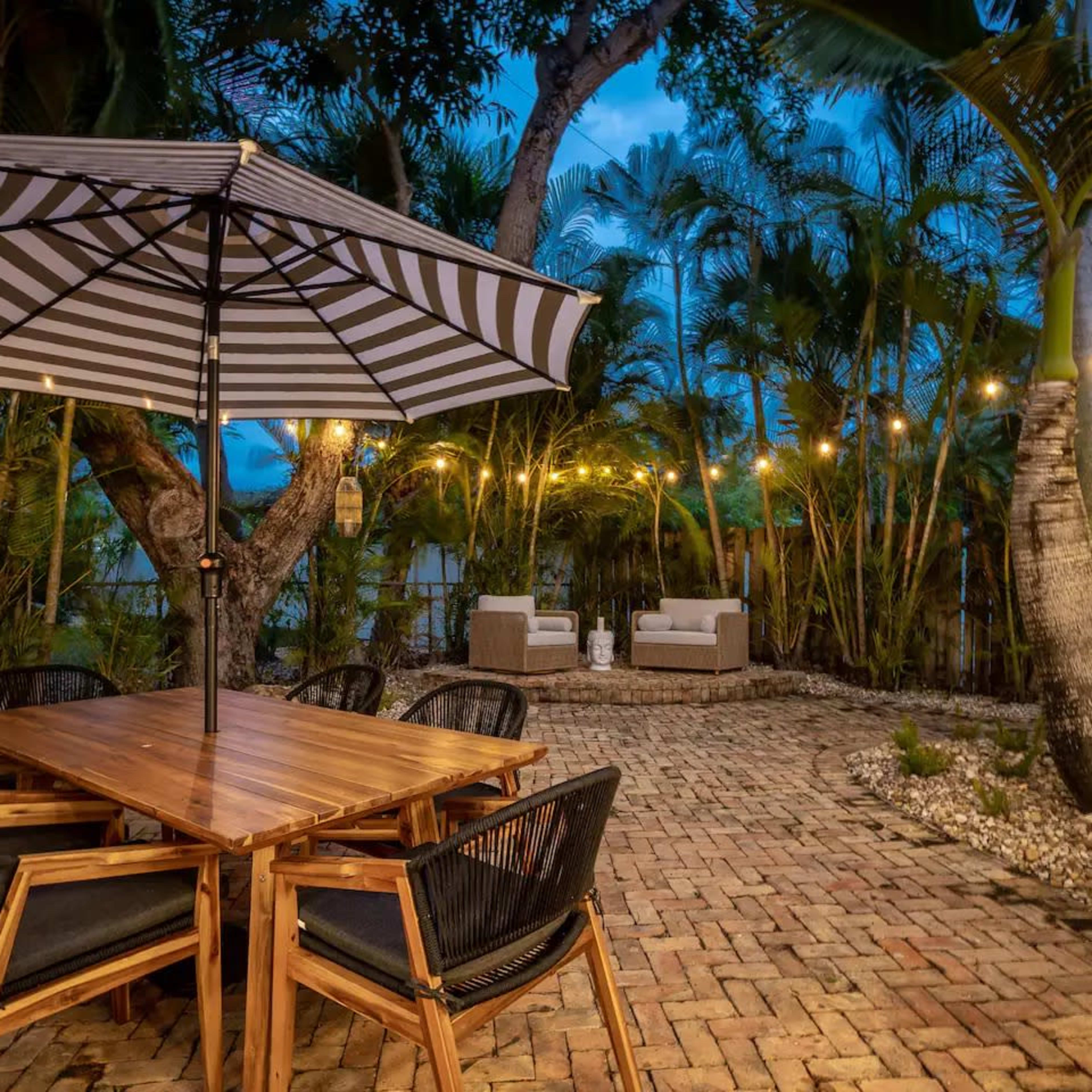 A wooden dining table with black chairs sits under a striped umbrella in a landscaped outdoor space, adorned with string lights and surrounded by palm trees.