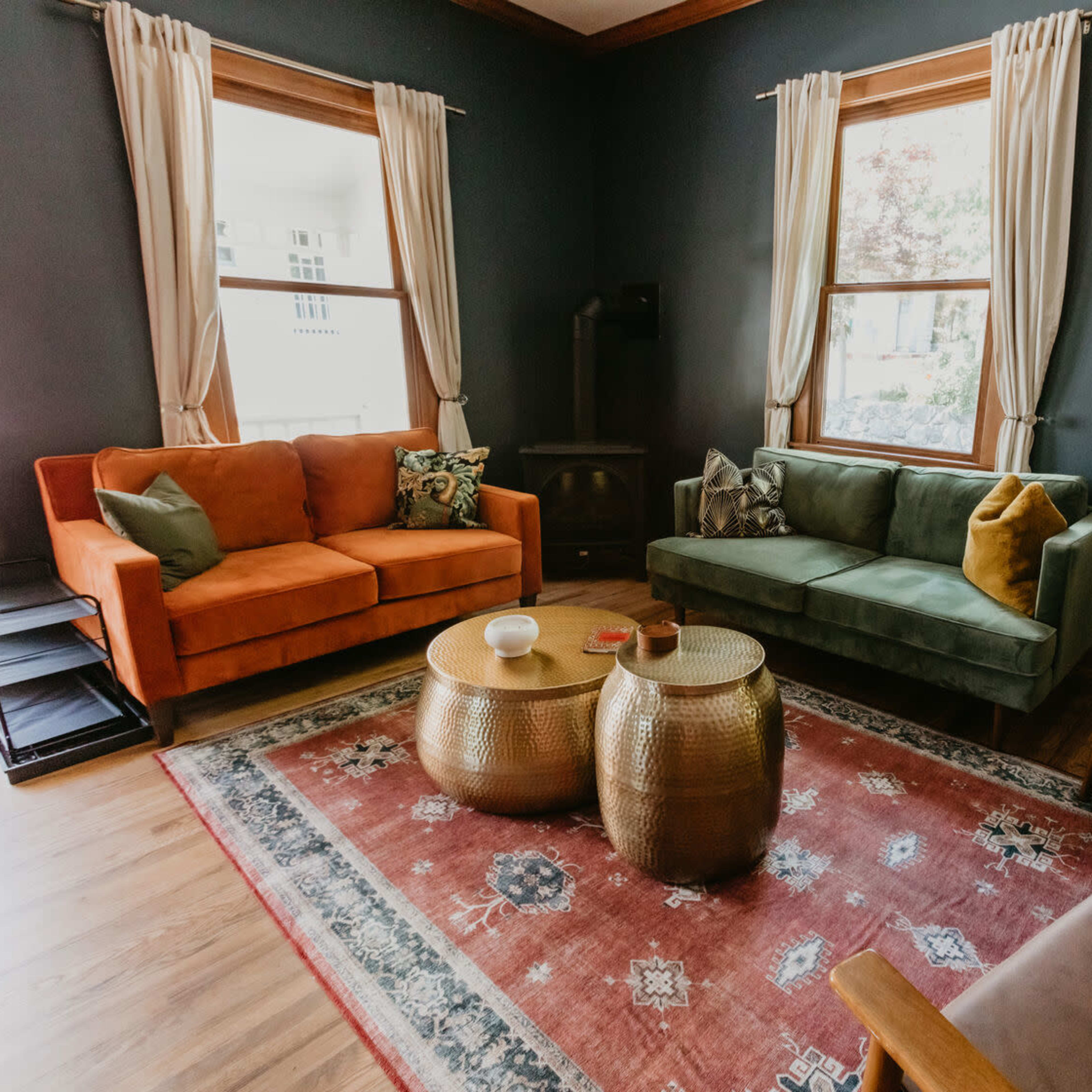 The living room features two sofas in orange and green, a patterned rug, and two round metal coffee tables against a dark wall.