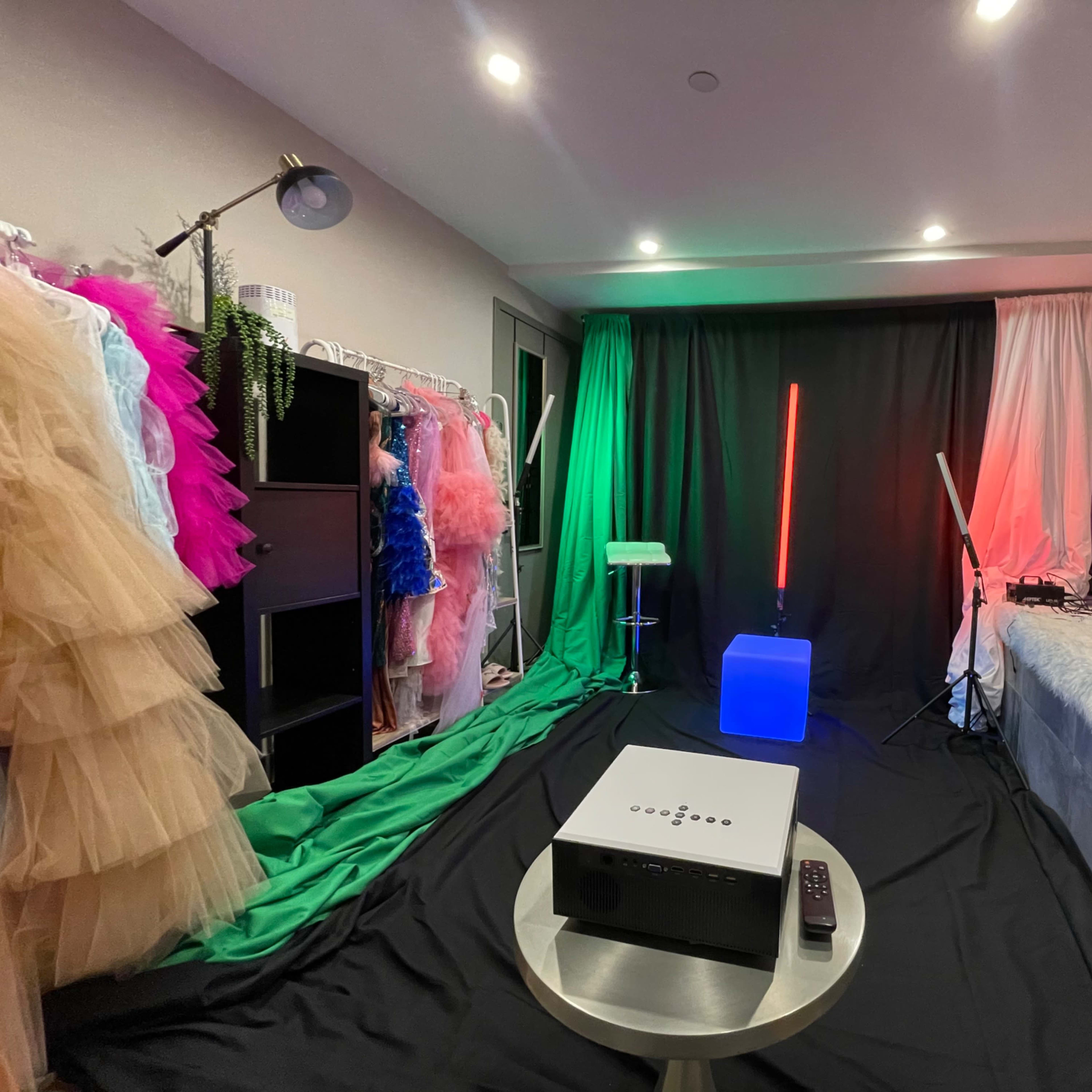 A room set up for a photoshoot, featuring colorful dresses on a rack, a projector on a table, and various lighting equipment.