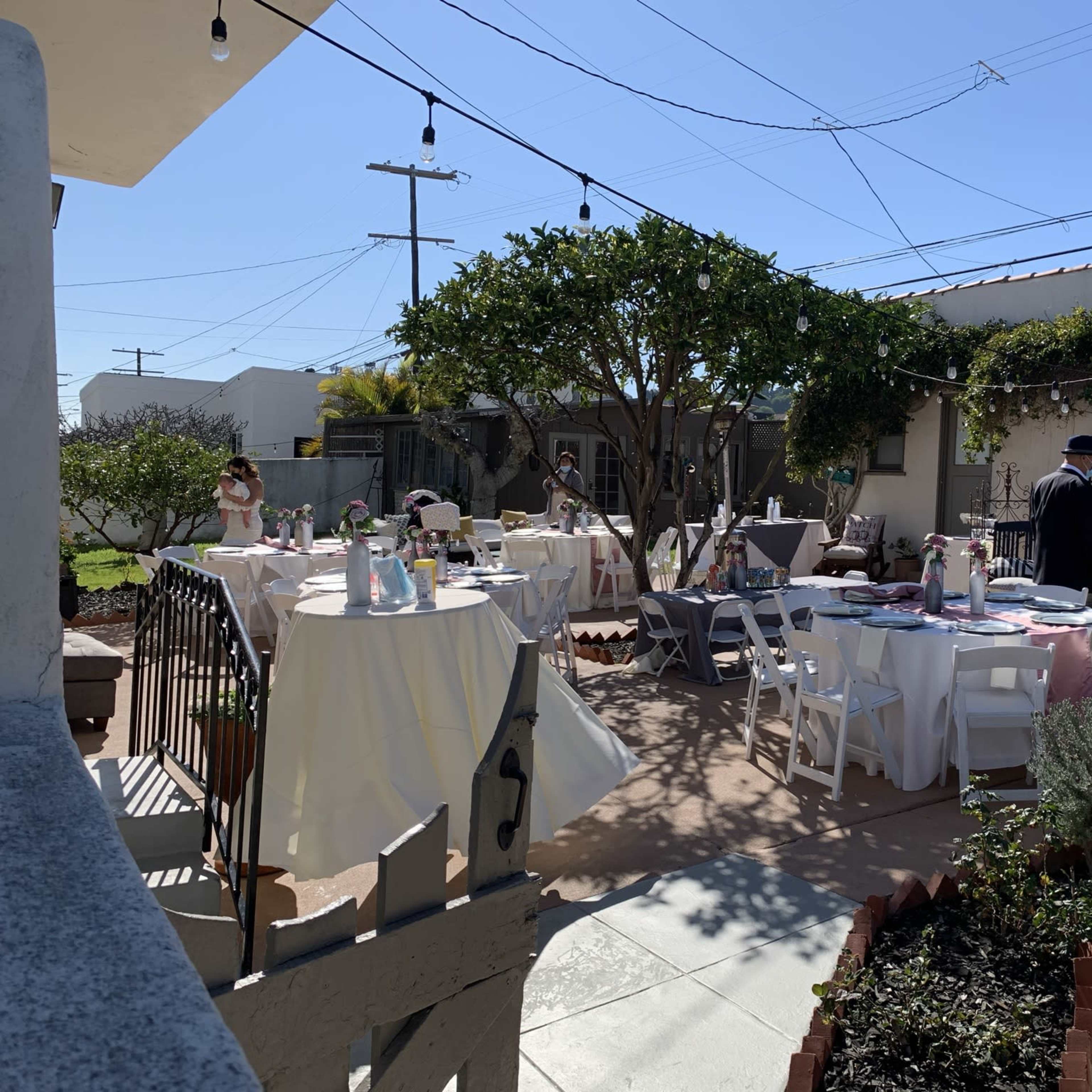 An outdoor dining area set up with tables and chairs under clear blue skies, surrounded by greenery.