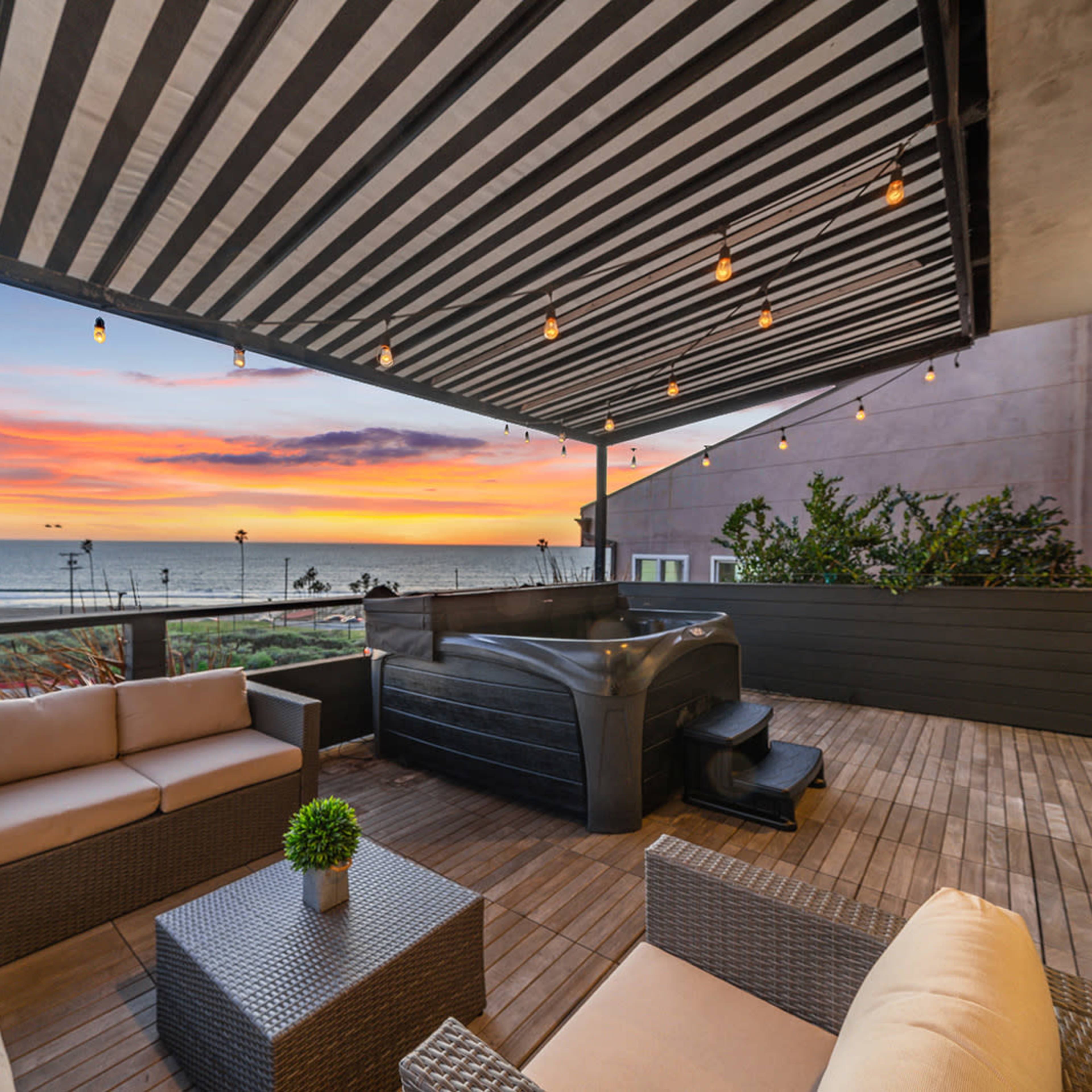 A patio with striped awnings features a hot tub and comfortable seating, overlooking a sunset view of the ocean.